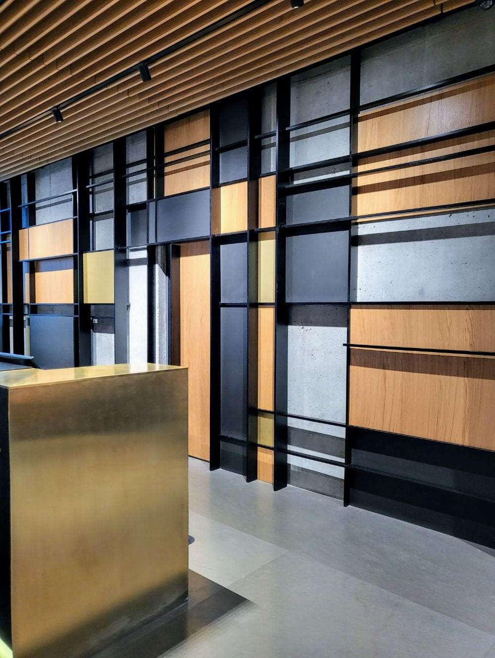 Abstrakcyjna aranżacja ściany z elementami drewna, betonu i czarnych metalowych profili, uzupełniona złotą ladą recepcyjną w nowoczesnym wnętrzu.