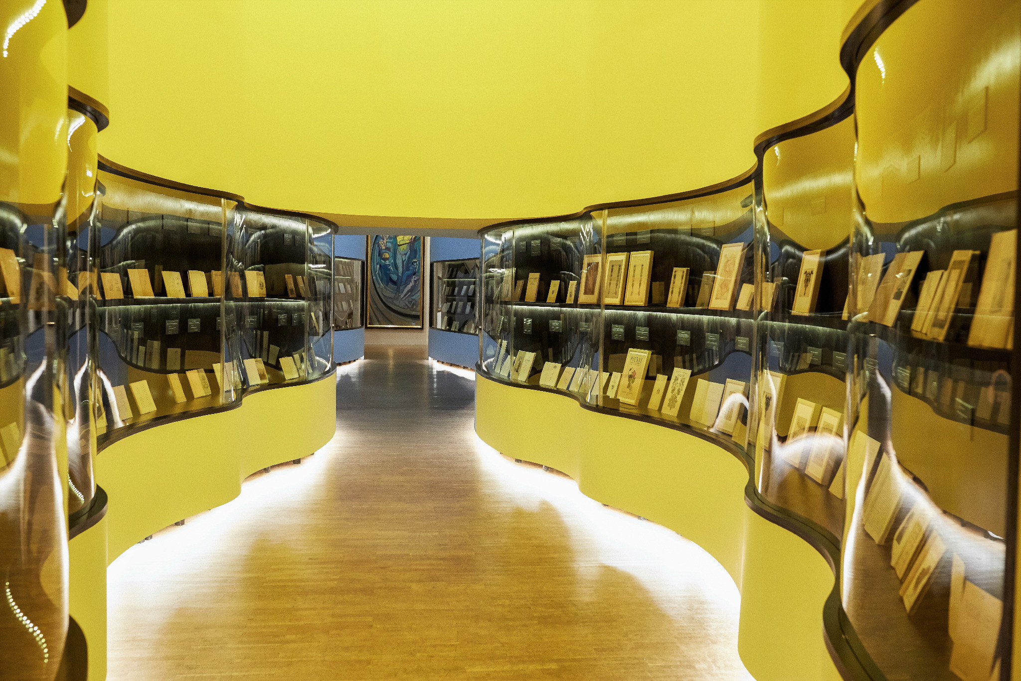 Zakrzywiona, przeszklona witryna ekspozycyjna z żółtymi elementami, prezentująca książki i dokumenty w oświetlonym wnętrzu z żółtym sufitem.
