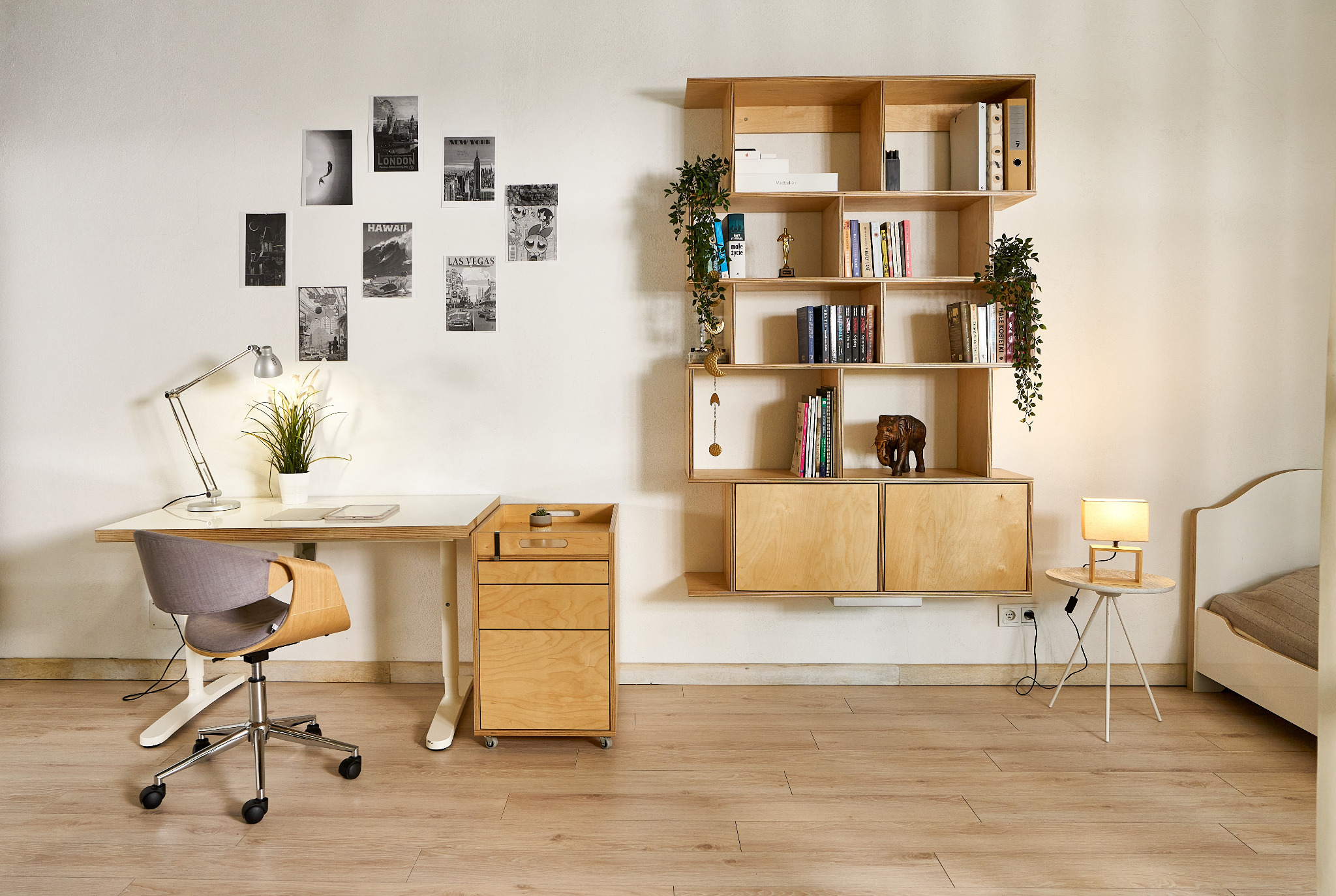 Jasne biuro z drewnianym biurkiem, krzesłem na kółkach, kontenerkiem z szufladami i wiszącą półką na książki z roślinami, uzupełnione lampką na biurku i stoliku nocnym oraz plakatami na ścianie.