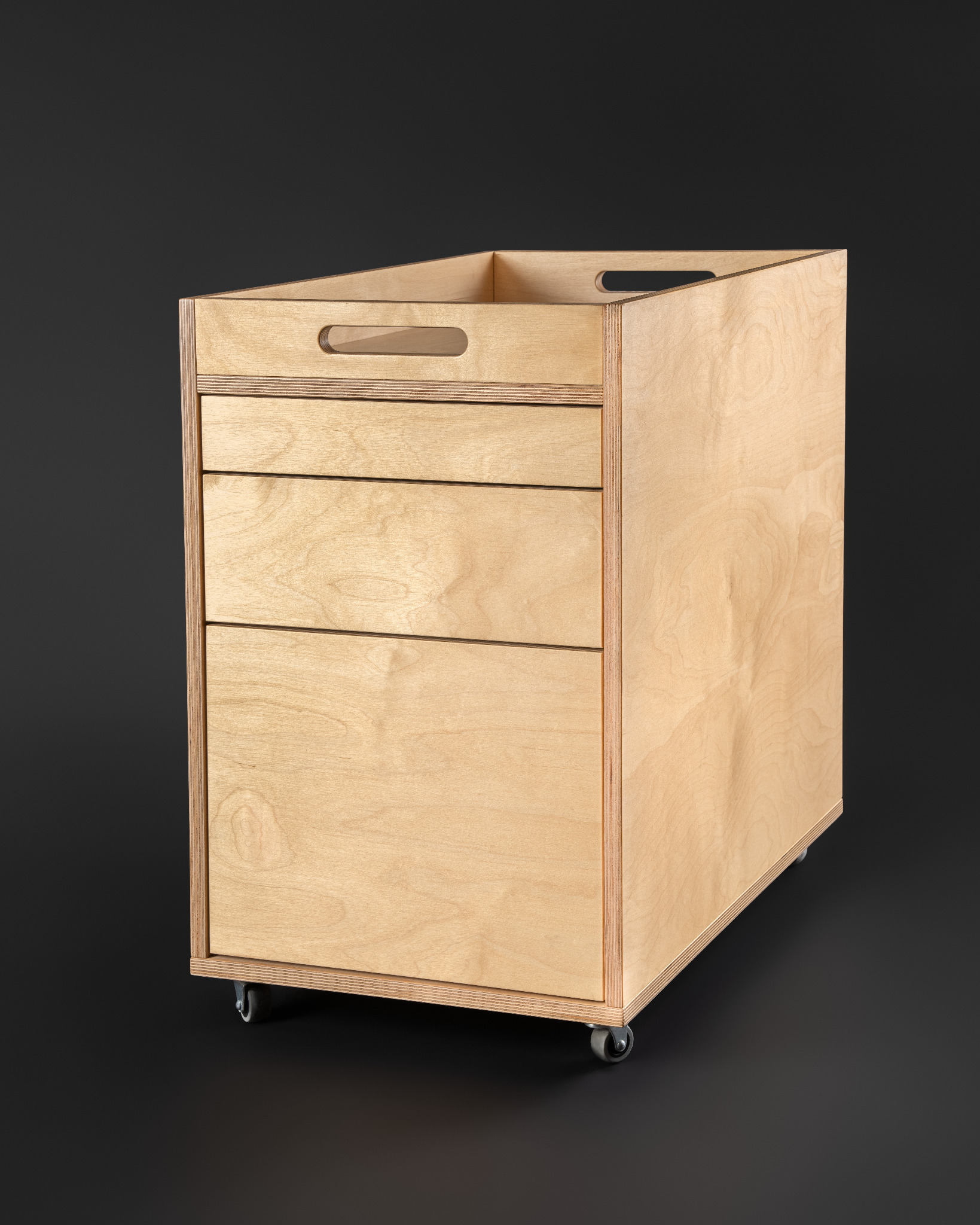 Mobilna szafka z trzema szufladami wykonana ze sklejki, z uchwytami wyciętymi w górnej części, na kółkach, na ciemnym tle.
