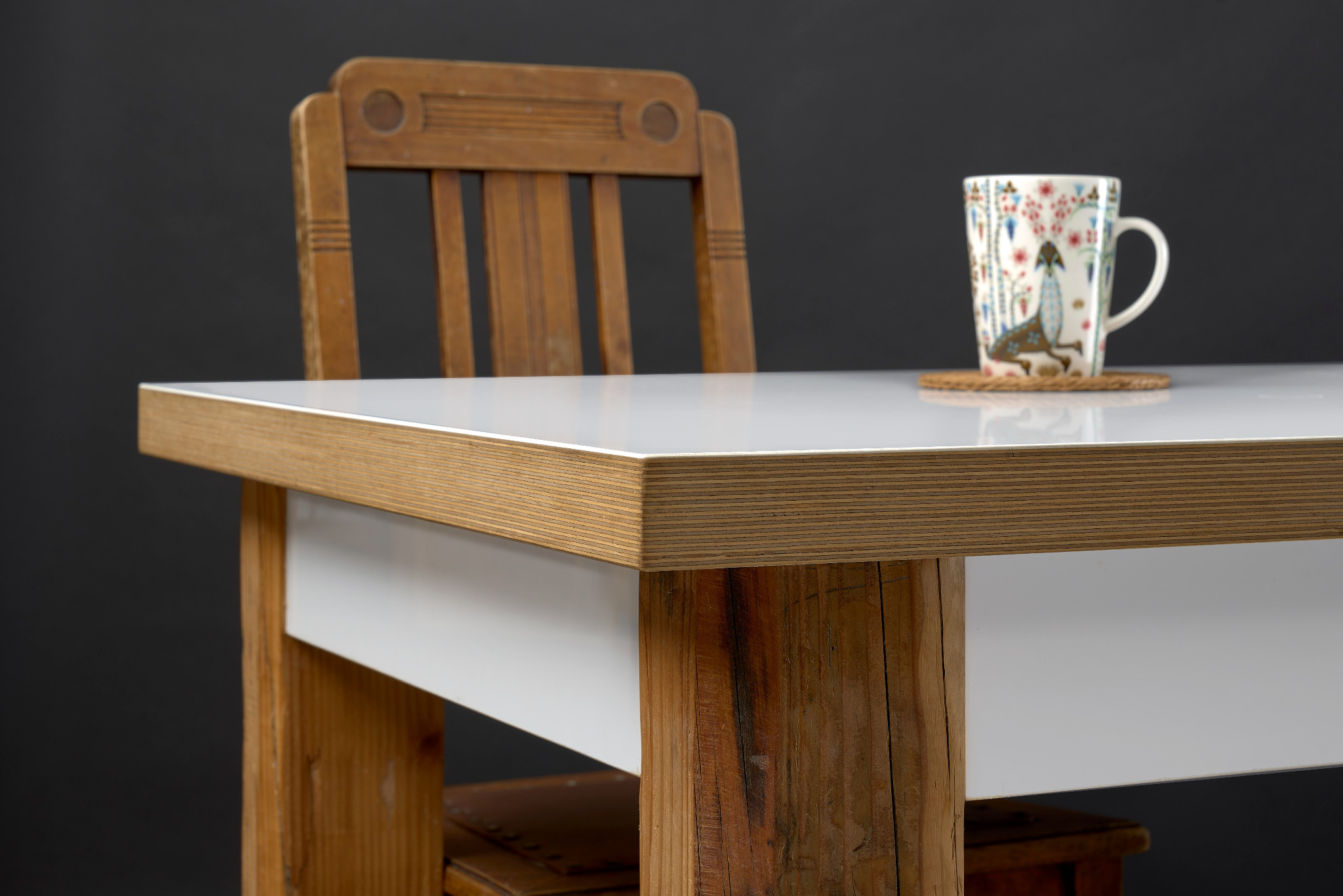 Biały blat stołu z drewnianym obramowaniem i nogami z surowego drewna, w tle krzesło z drewna i kubek z wzorem na podkładce.
