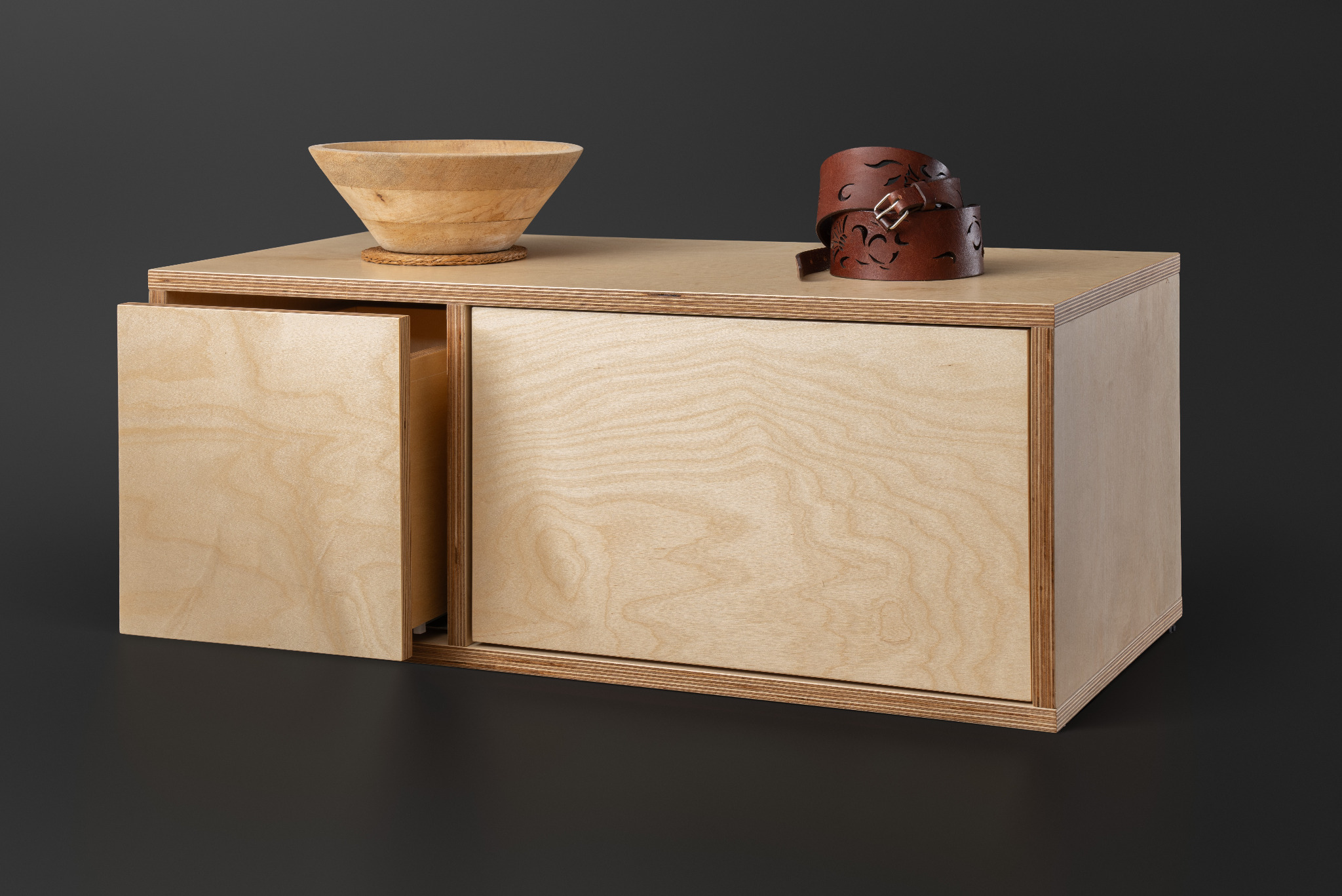 Niska komoda z jasnej sklejki z wysuwanymi drzwiczkami, na blacie drewniana misa i skórzany pasek z wycinanym wzorem, na ciemnoszarym tle.