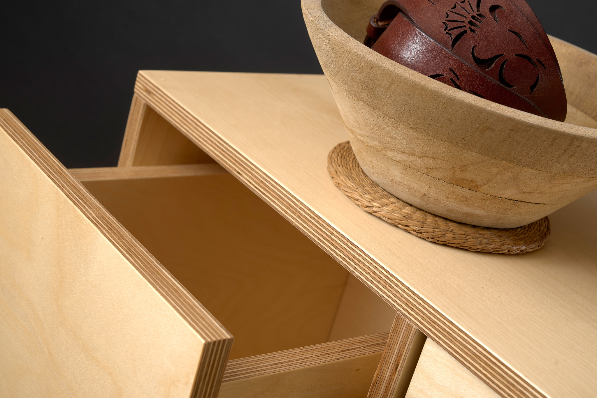 Szafka z jasnej sklejki z wysuwanymi szufladami, na blacie drewniana misa z brązowym paskiem skórzanym z wycinanym wzorem.