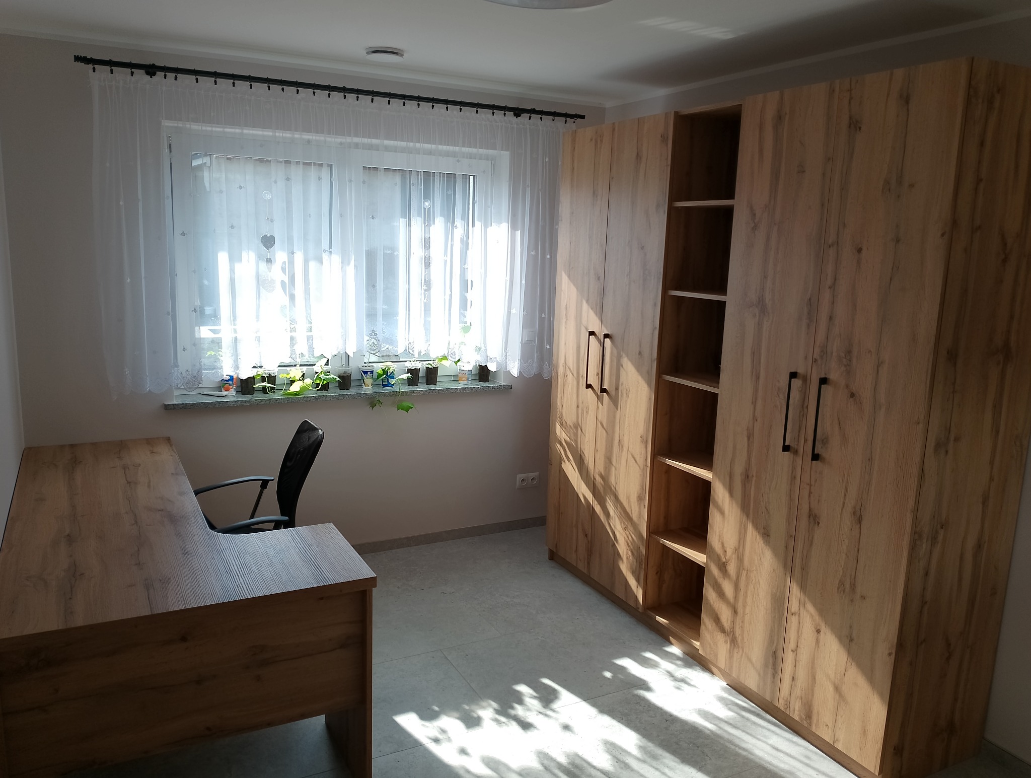 Wnętrze pokoju z biurkiem narożnym i wysoką szafą wnękową z półkami, wykonane z drewna w odcieniach brązu, oświetlone naturalnym światłem wpadającym przez okno z firanką i roślinami.