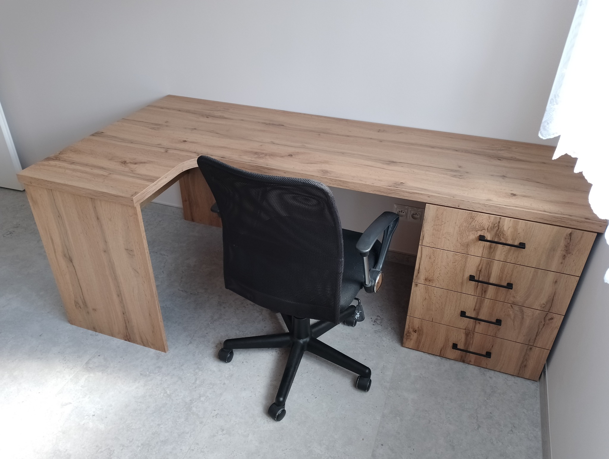 Biurko narożne z drewnopodobnym blatem i trzema szufladami po prawej stronie, z czarnym krzesłem biurowym na kółkach ustawionym przed nim.