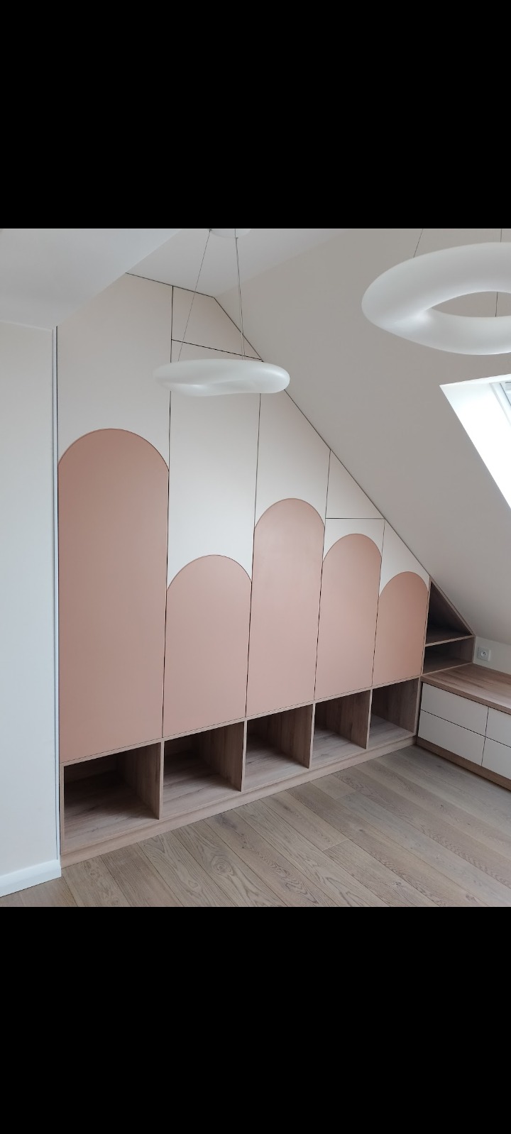 Zabudowa pod skosem z szafkami z jasnego drewna i różowymi, zaokrąglonymi frontami, oświetlona nowoczesnymi lampami wiszącymi