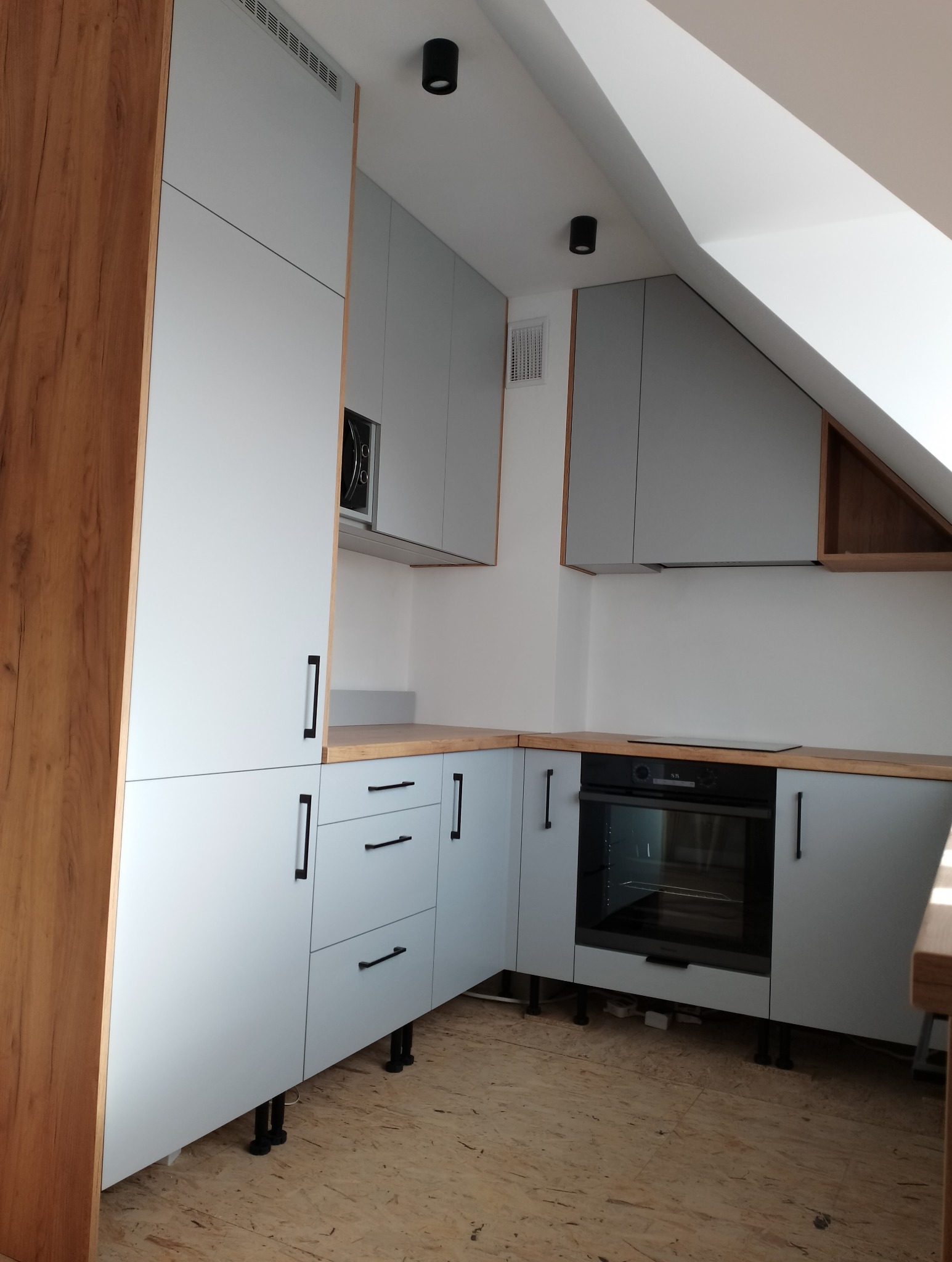 Jasnoszara zabudowa kuchenna z drewnianymi akcentami w skosach, czarne podłużne uchwyty, piekarnik i mikrofalówka w zabudowie, czarne punktowe oświetlenie sufitowe.