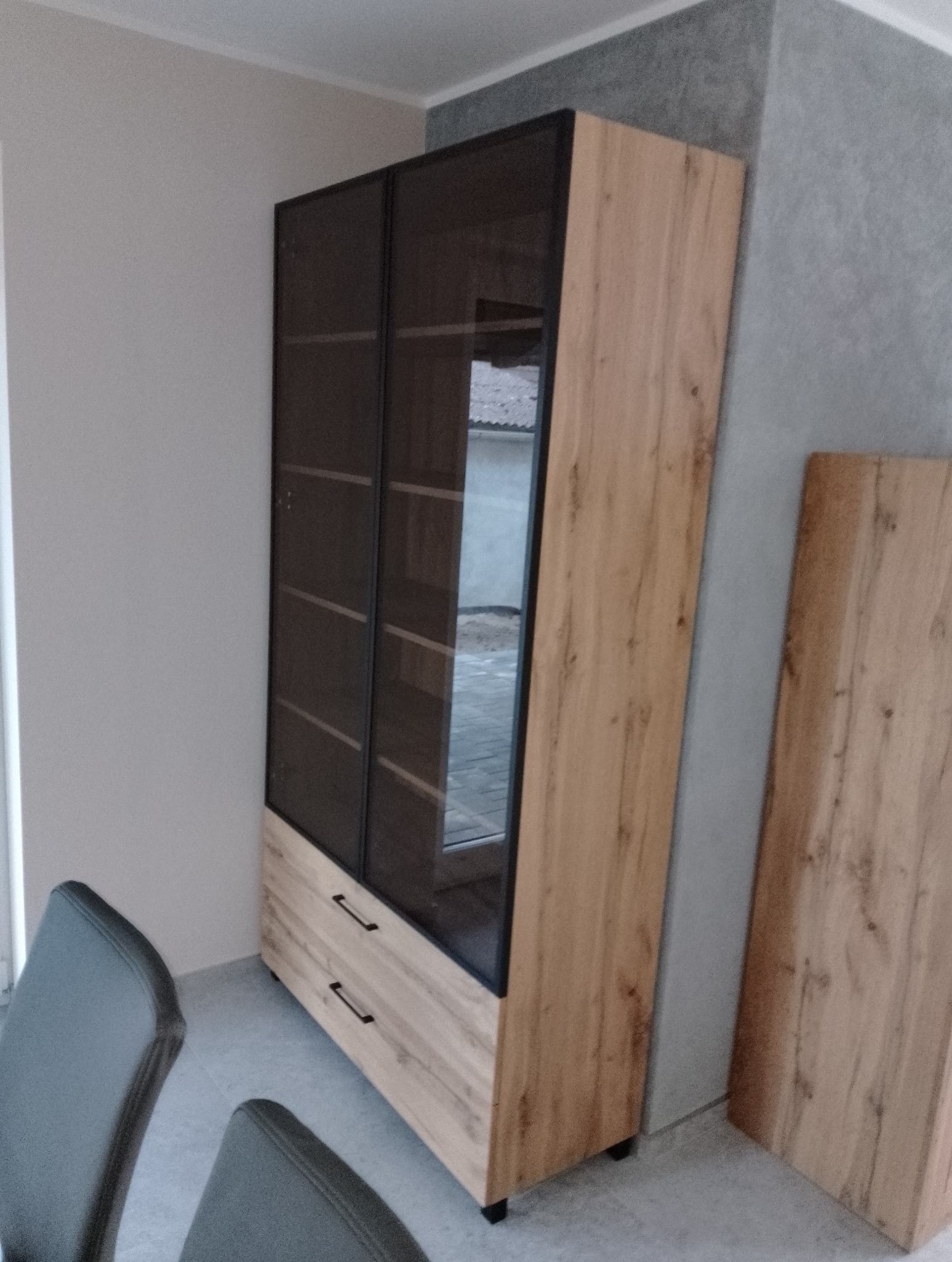 Dwudrzwiowa witryna z szufladami w okleinie drewnopodobnej z czarnymi ramami szklanych frontów, widok z boku, fragment krzesła w kadrze.