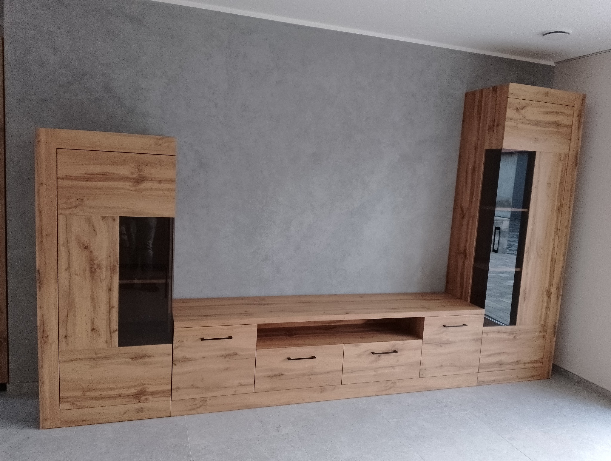 Jasne, drewnopodobne meble modułowe z ciemnymi uchwytami, składające się z szafek, szuflad i przeszklonych witryn na tle szarej ściany.