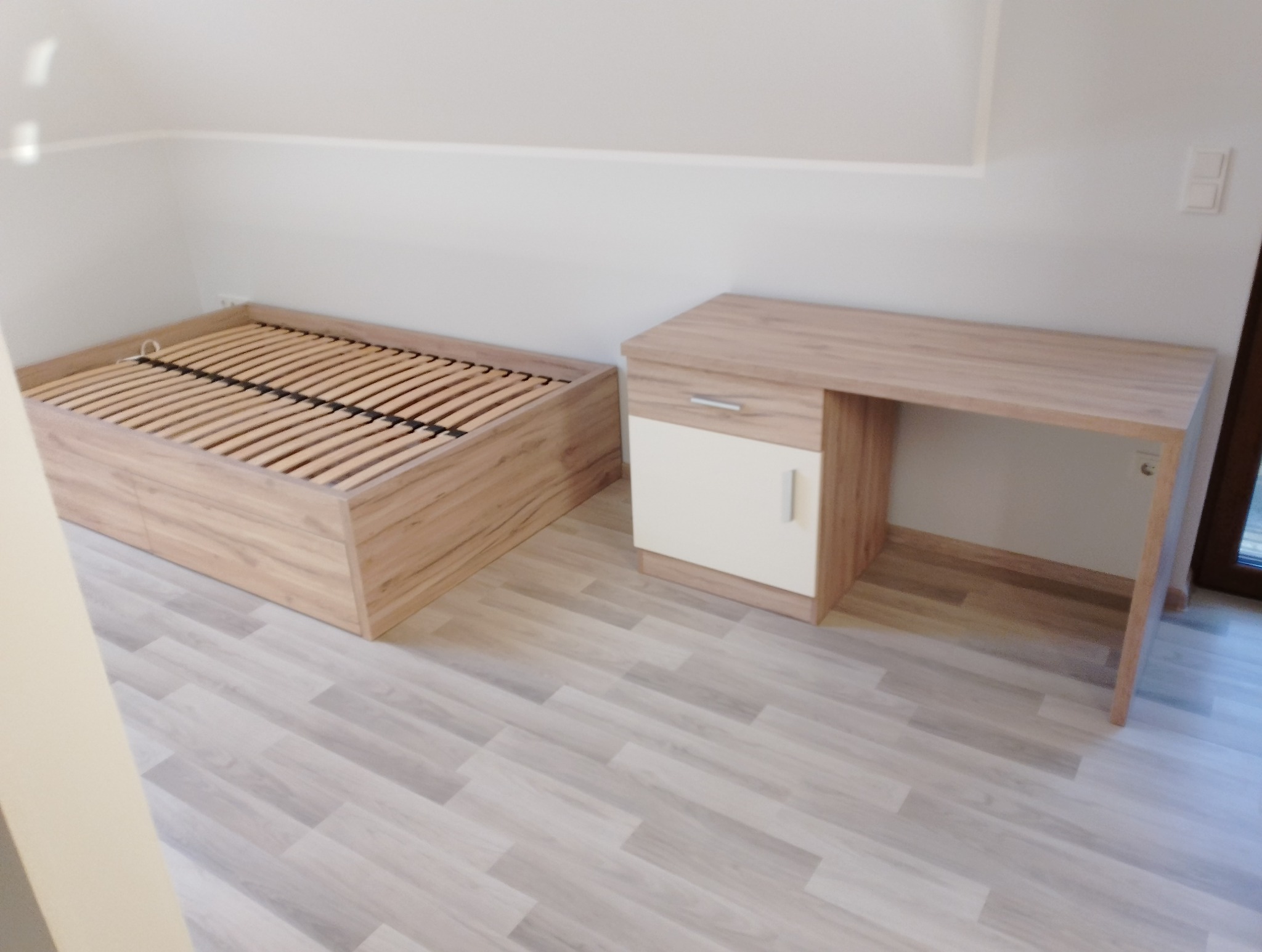 Rama łóżka z drewnianymi listwami i biurko z szafką w pokoju z szarą podłogą imitującą drewno.