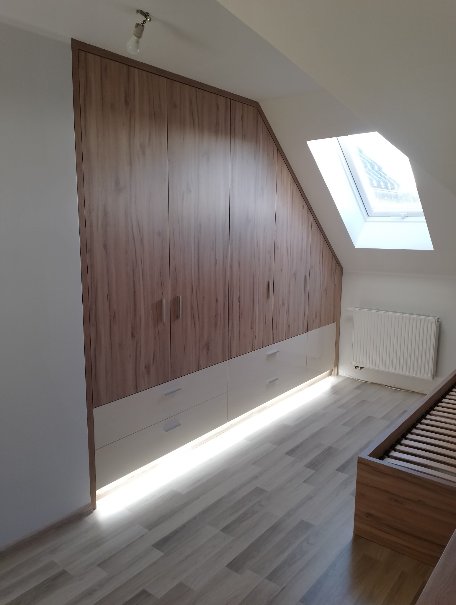 Szafa wnękowa z drewnopodobnymi frontami i białymi szufladami, oświetlenie LED u dołu, dopasowana do skosu poddasza, widok na okno dachowe i fragment łóżka.