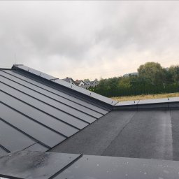 Przemysław Mikołajczak - Szary dach z blachy na tle zabudowań Ostrowa Wielkopolskiego w pochmurny dzień. Widoczne łączenia blachy i fragment dachu pokrytego papą.