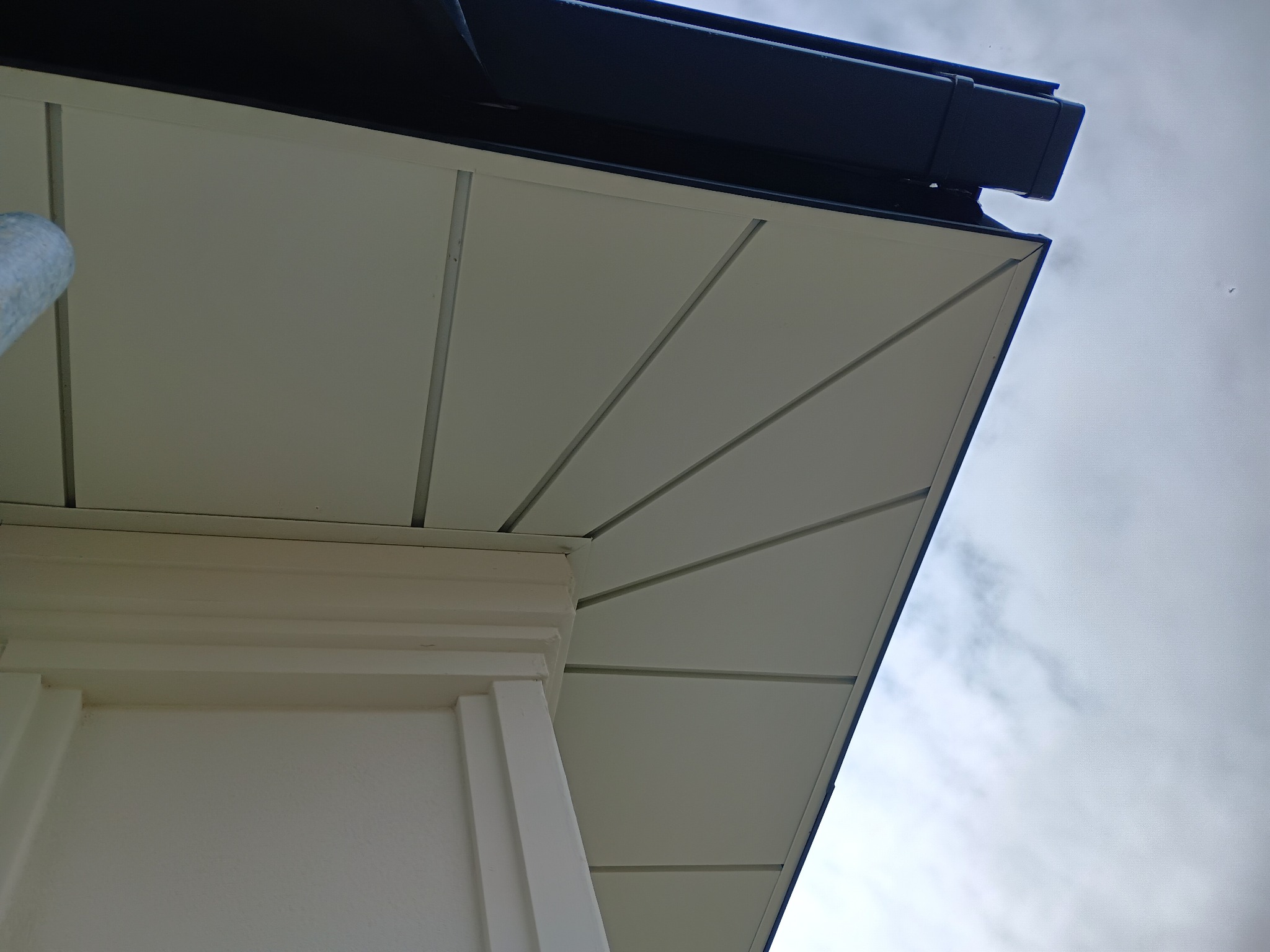 Szczegółowe ujęcie okapu dachowego z ciemną rynną, jasną podbitką i fragmentem ściany budynku w jasnej tonacji, sfotografowane pod kątem.