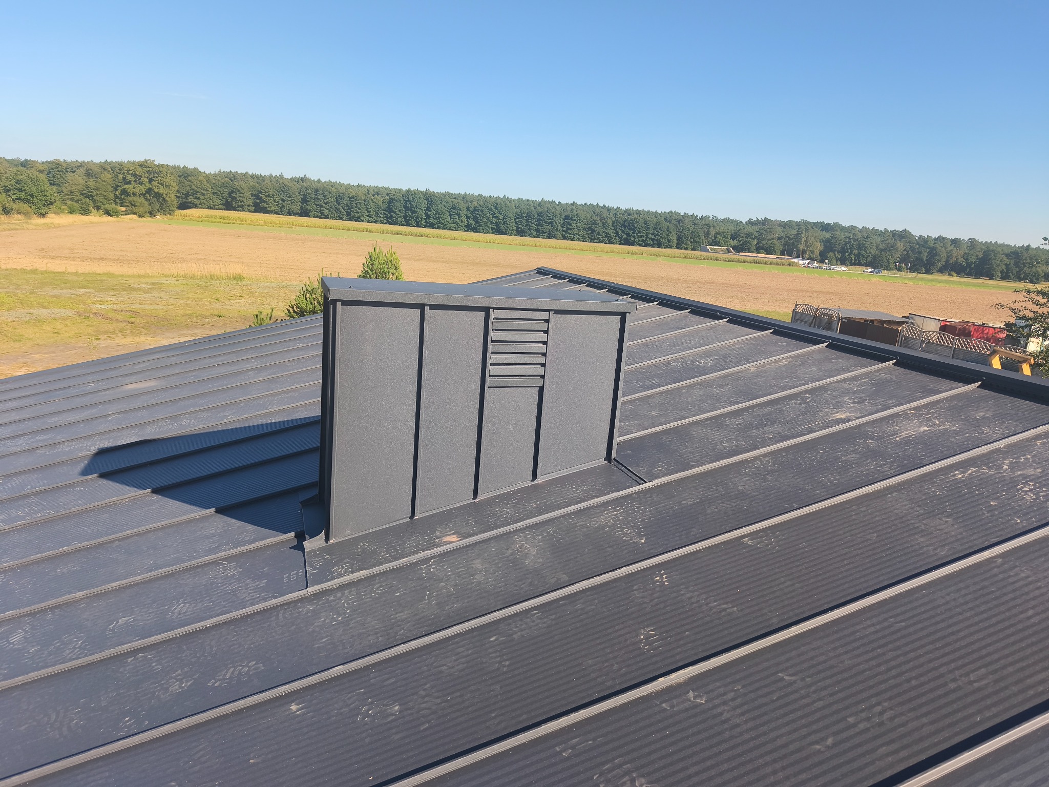 Szary dach z blachy na tle pola i lasu. Widoczny komin z wentylacją. Ujęcie z góry, prezentujące szczegóły pokrycia i obróbki blacharskiej.