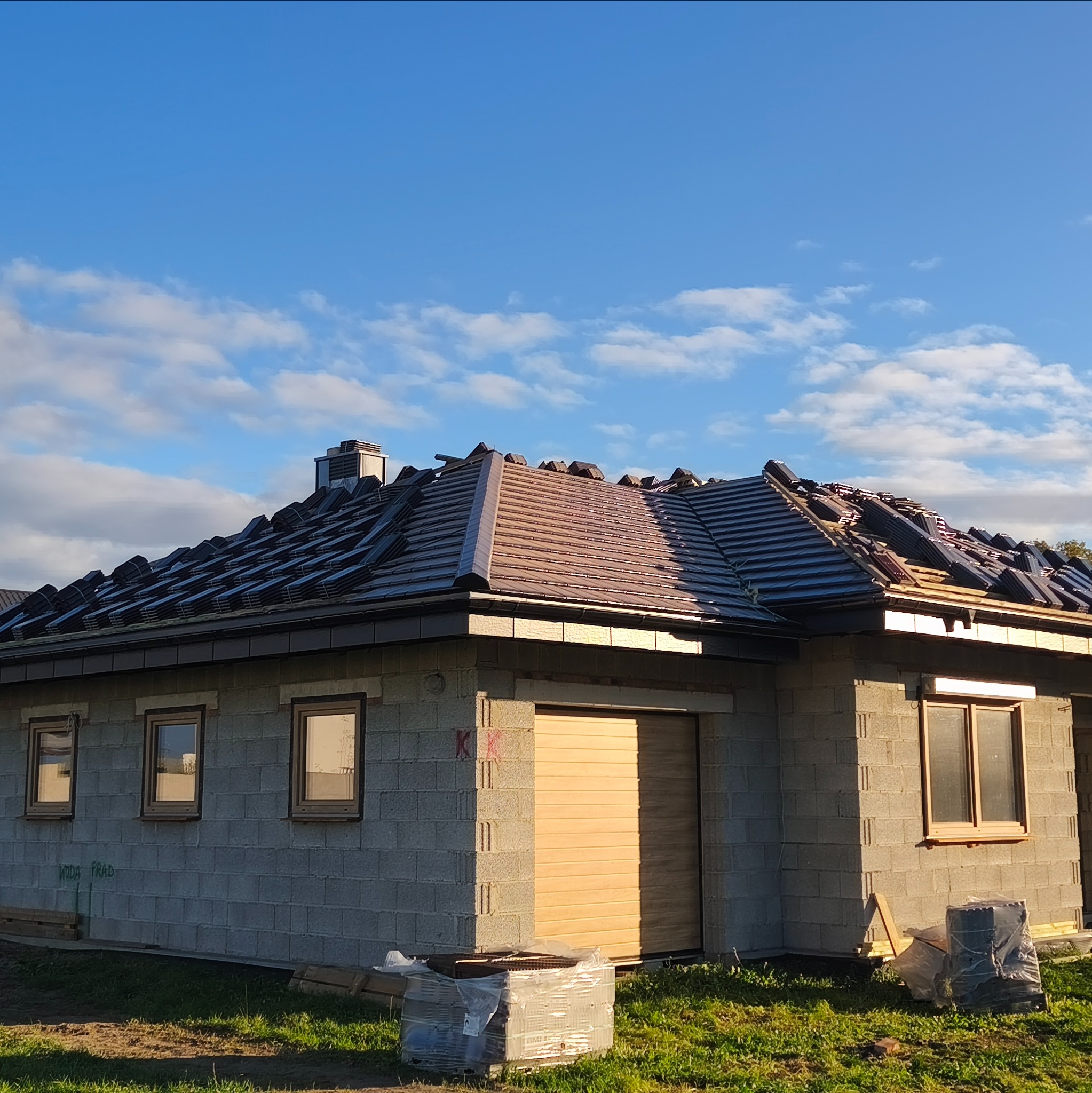 Dom w trakcie krycia dachu brązową dachówką ceramiczną. Widoczne ułożone dachówki i fragmenty więźby. Ściany budynku z szarego bloczka betonowego.