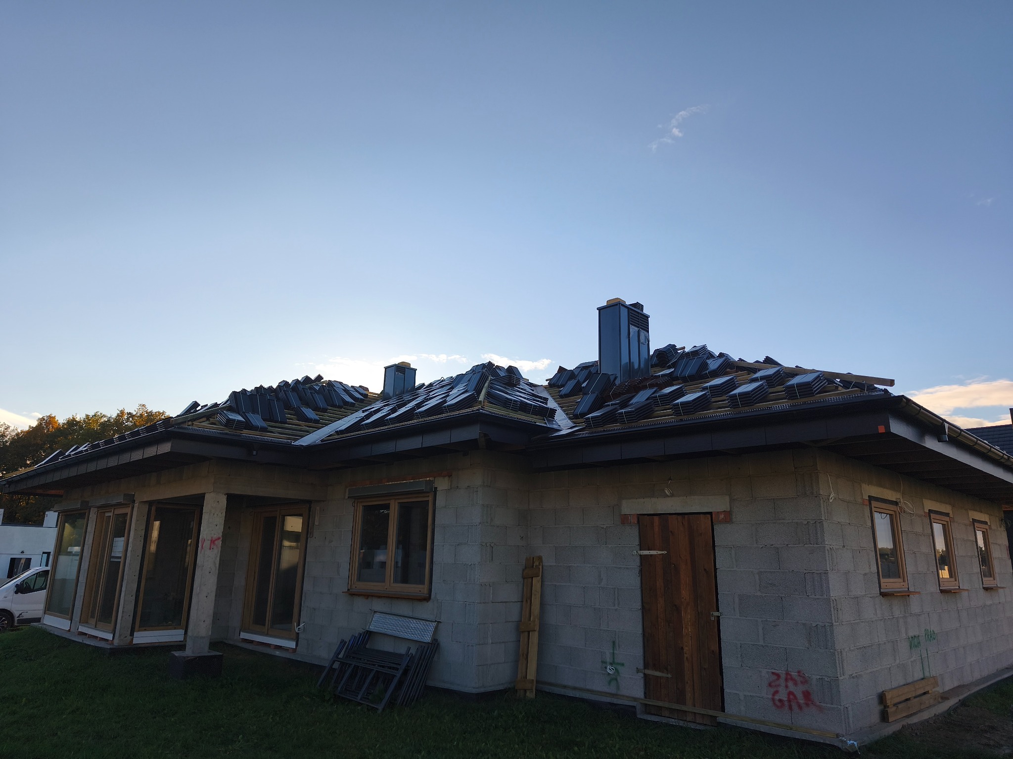 Dom w trakcie krycia dachu dachówką ceramiczną, widoczne kominy i fragment nieba. Budynek z szarego bloczka, okna w drewnianych ramach, częściowo zamontowane.