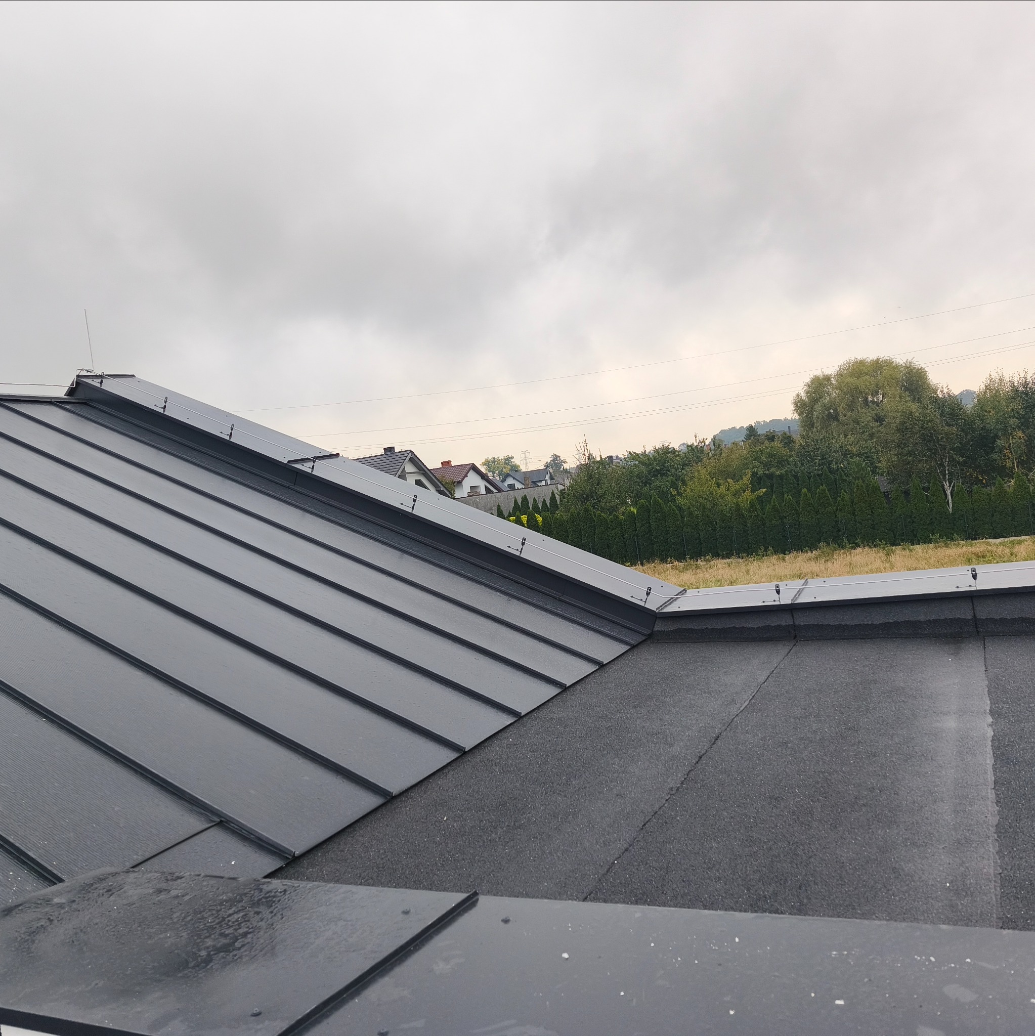 Szary dach z blachy na tle zabudowań Ostrowa Wielkopolskiego w pochmurny dzień. Widoczne łączenia blachy i fragment dachu pokrytego papą.
