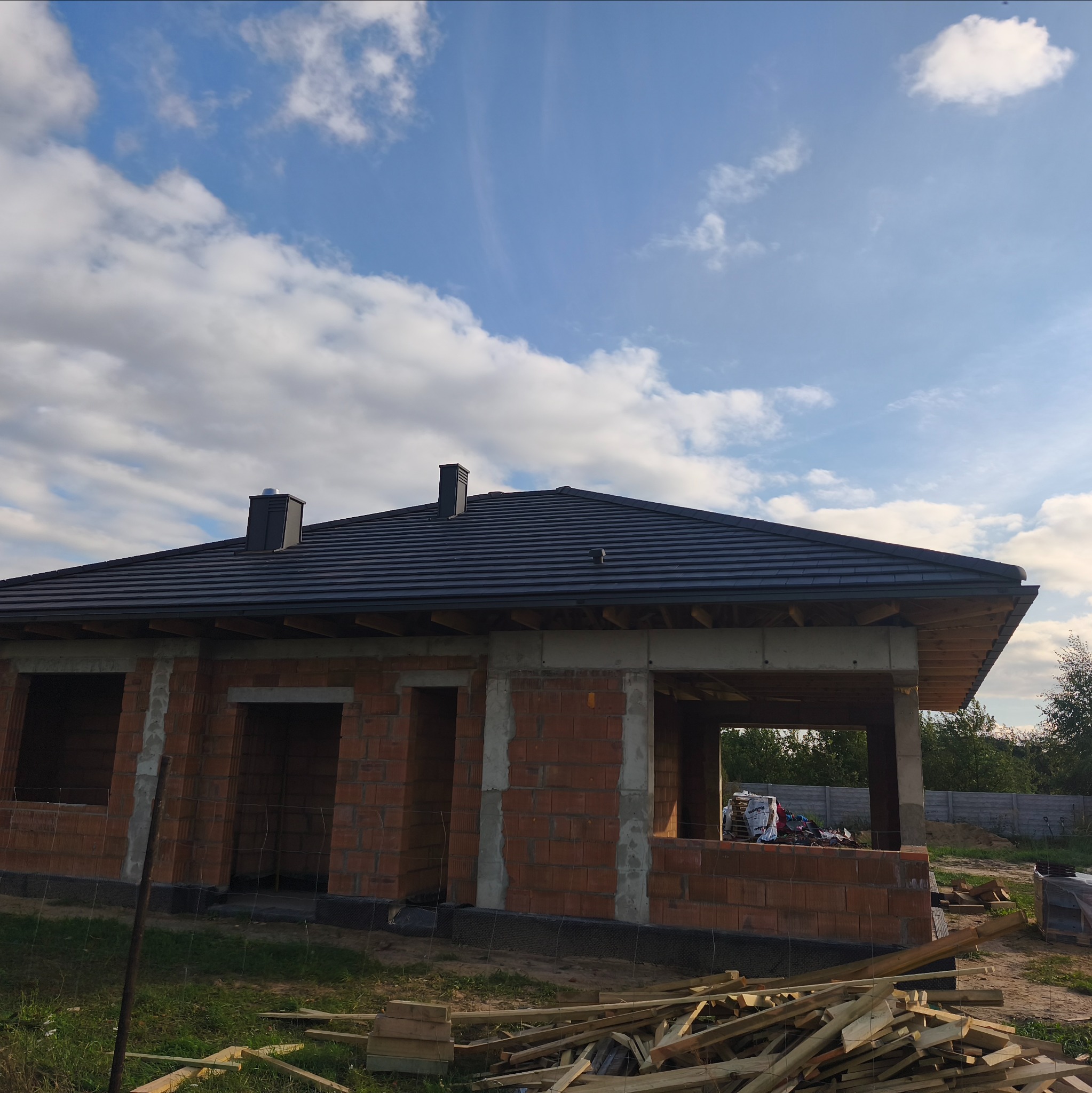 Dom w trakcie budowy z ciemnym dachem z dachówki ceramicznej. Widoczne ceglane ściany i drewniane elementy konstrukcyjne. Błękitne niebo z chmurami w tle.
