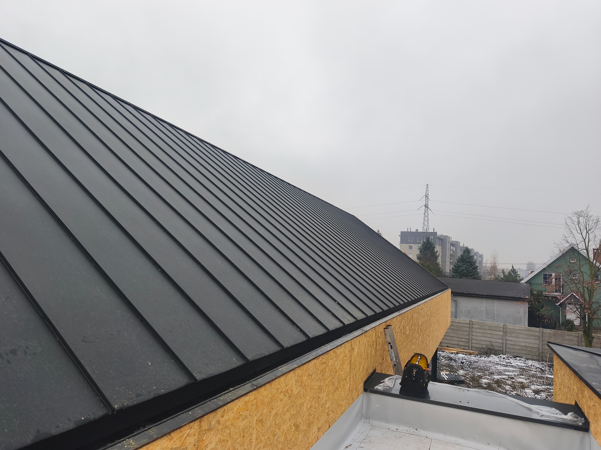 Nowoczesny dach z blachy na budynku w trakcie budowy, widok z góry, z elementami konstrukcyjnymi i narzędziami dekarskimi. Tło stanowią budynki mieszkalne i pochmurne niebo.