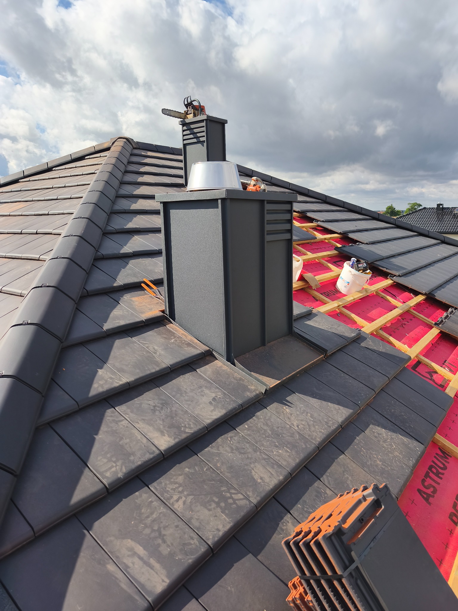 Częściowo pokryty dach z ciemnej dachówki ceramicznej, widoczny komin z piłą łańcuchową na szczycie, fragment więźby i membrany dachowej. Ujęcie z góry.