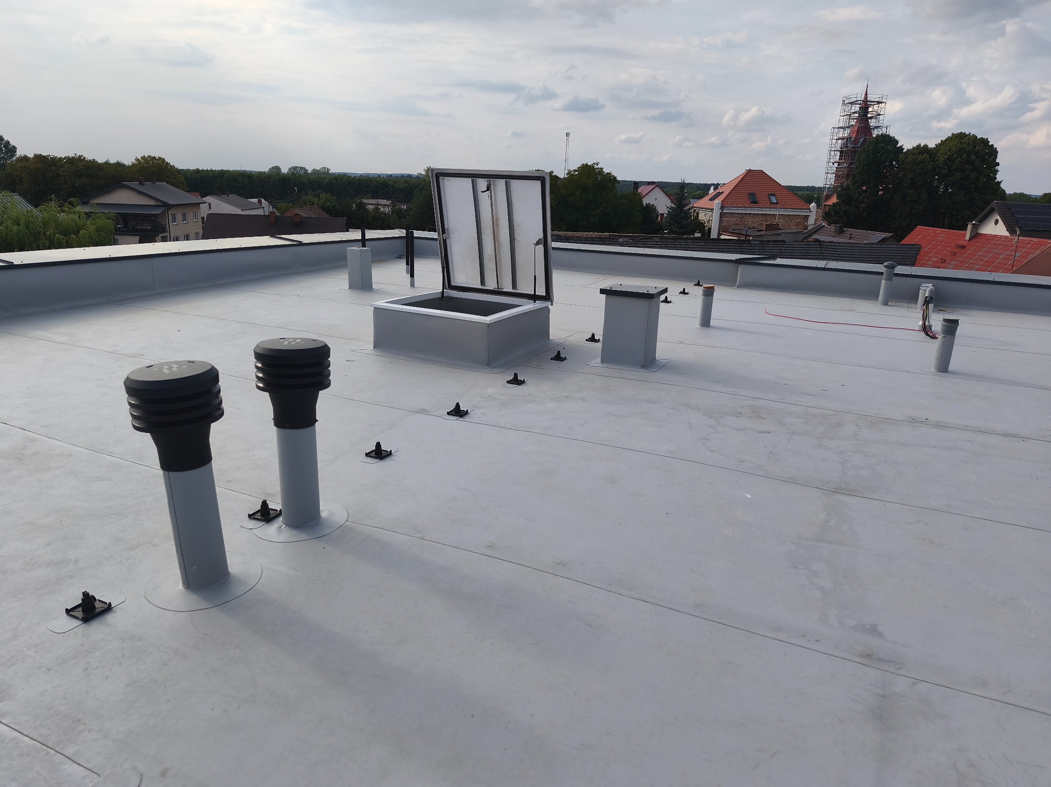 Jasnoszary dach płaski z wywietrznikami i wyłazem dachowym. W tle zabudowa miejska i kościół w rusztowaniach.