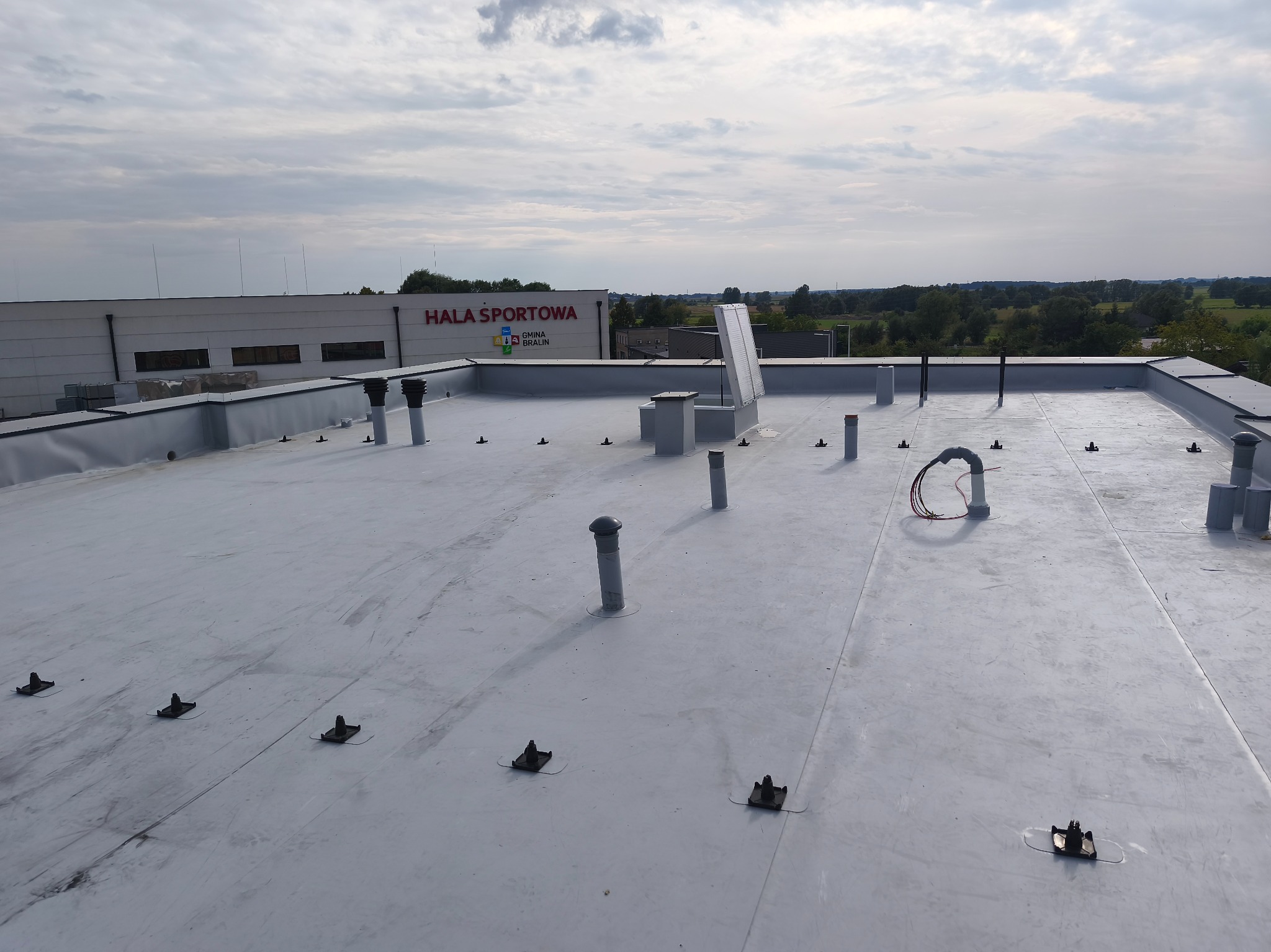 Szary, płaski dach hali sportowej z licznymi kominkami wentylacyjnymi i mocowaniami. W tle budynek hali i pochmurne niebo.