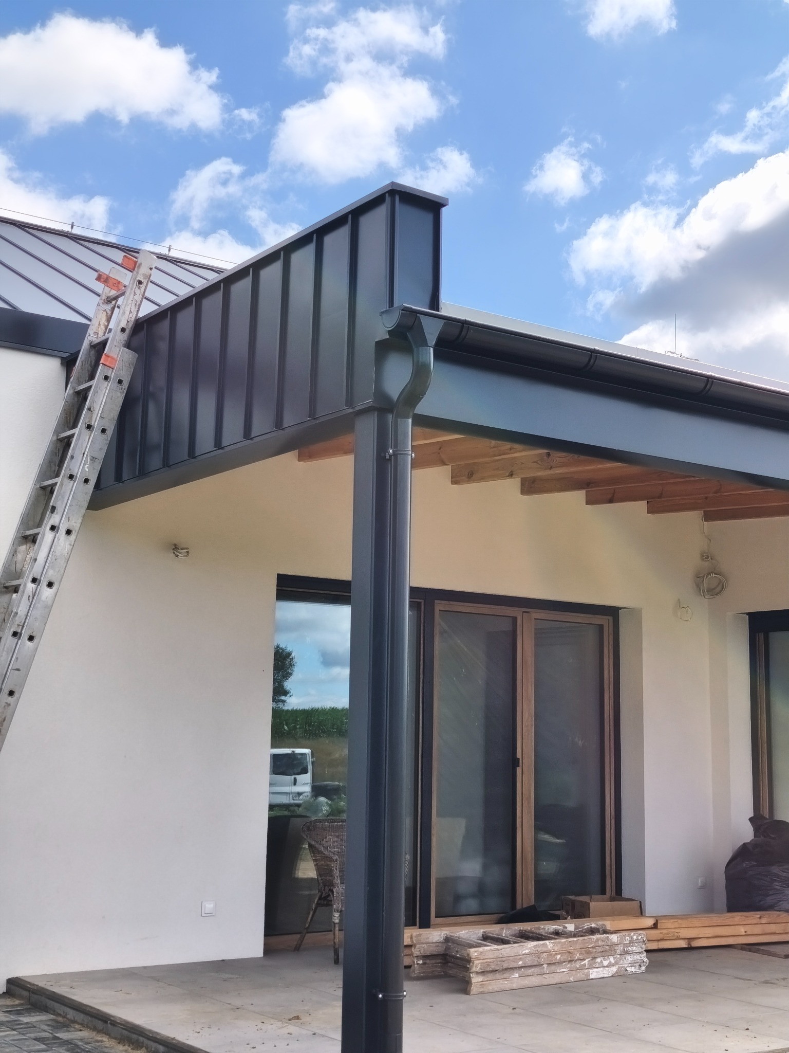 Nowoczesny dach z blachy na domu jednorodzinnym, z zamontowanymi rynnami w kolorze grafitowym. Aluminiowa drabina oparta o ścianę. Błękitne niebo w tle.