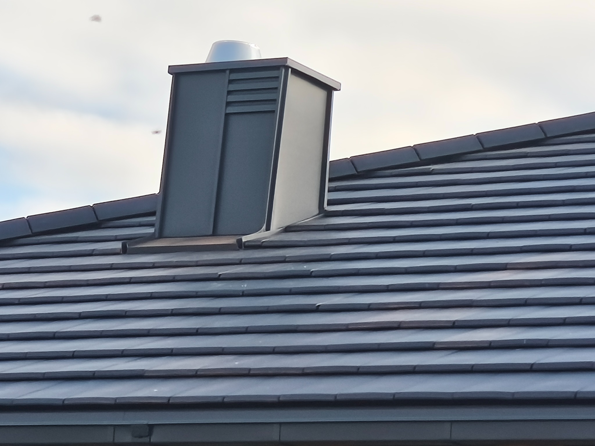 Grafitowy komin wentylacyjny na dachu z ciemnej dachówki ceramicznej, widok z dołu. Nowoczesne wykończenie dachu w spokojnej tonacji kolorystycznej.