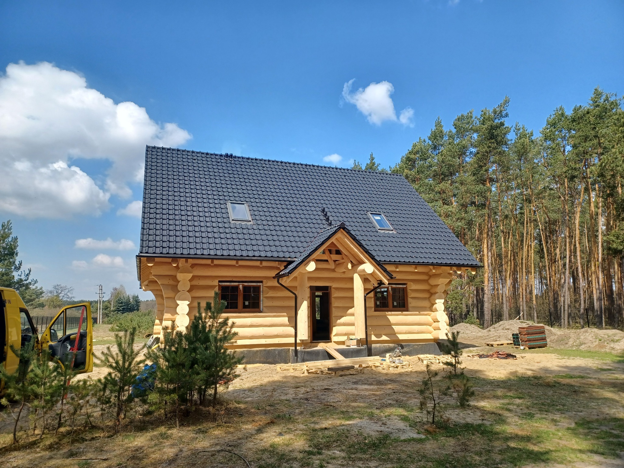 Nowoczesny dom z bali z ciemną dachówką, widoczny na tle lasu i błękitnego nieba. Elementy wykończeniowe w kolorze brązowym. Na pierwszym planie młode drzewka.