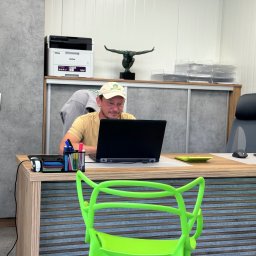 Travoni Usługi i Centrum Ogrodnicze - Mężczyzna w czapce z logo siedzi przy biurku z laptopem w biurze, widoczny drukarka, rzeźba i neonowe zielone krzesło.