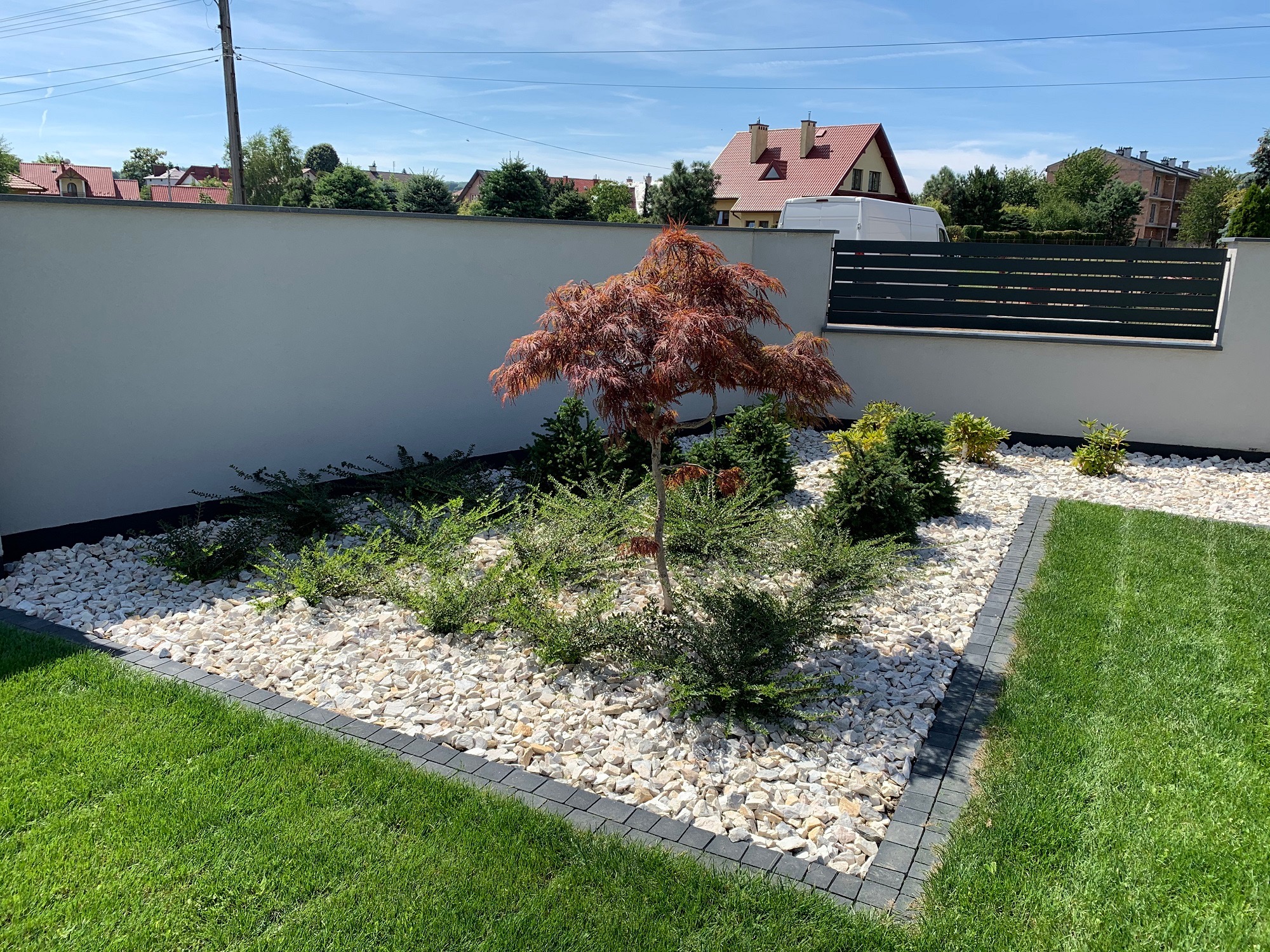 Kwadratowa rabata z białych kamieni obramowana szarą kostką, centralnie posadzony niewielki klon palmowy o bordowych liściach otoczony zielonymi krzewami, na pierwszym planie trawnik, w tle biały...