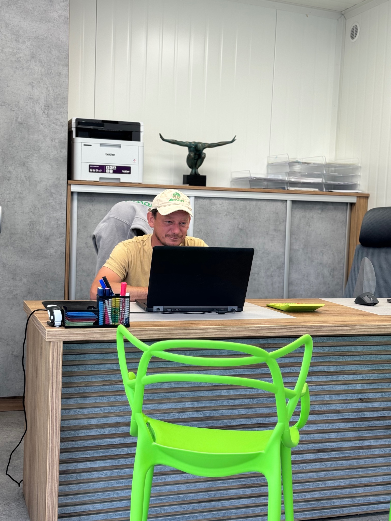 Mężczyzna w czapce z logo siedzi przy biurku z laptopem w biurze, widoczny drukarka, rzeźba i neonowe zielone krzesło.