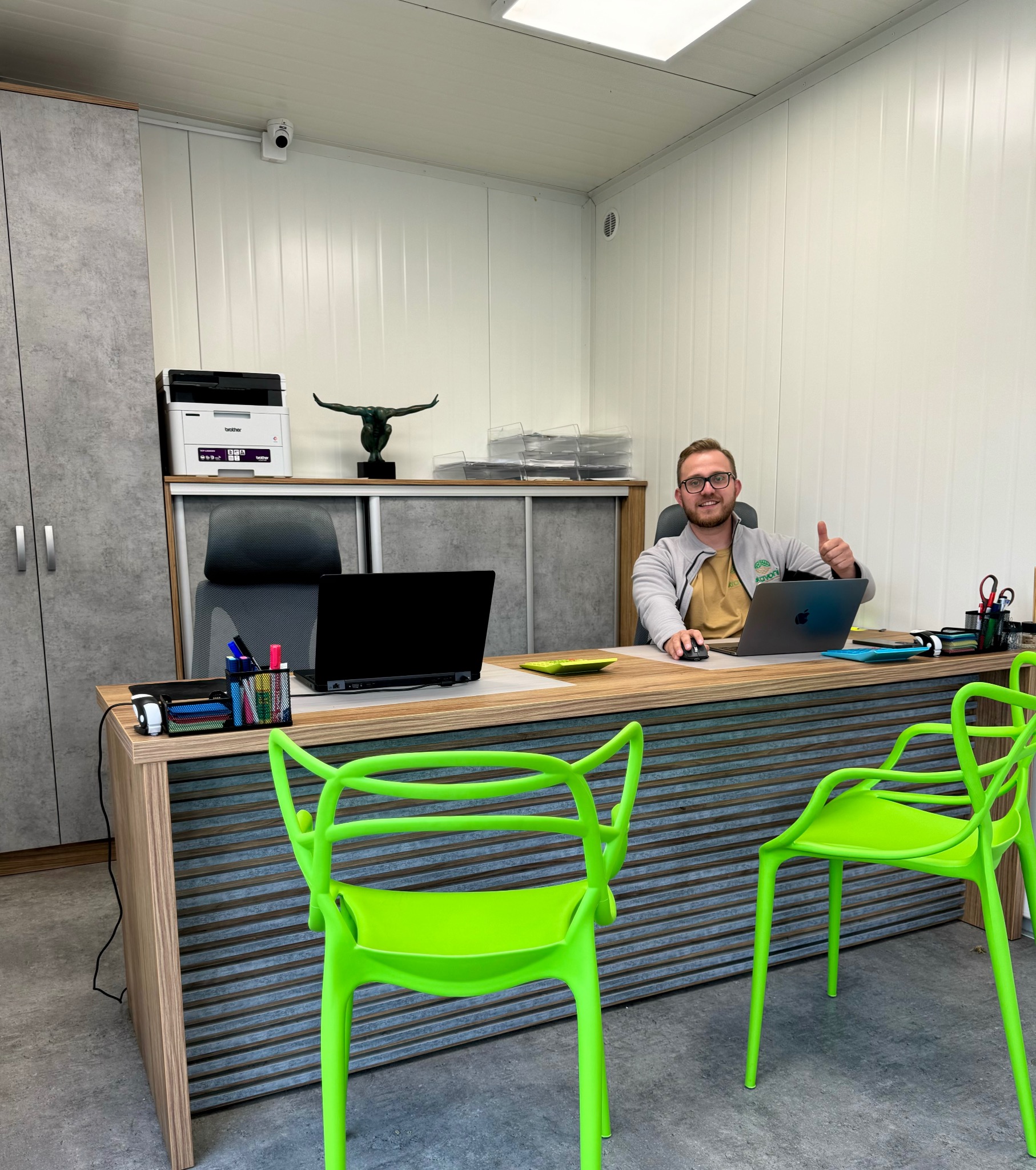 Mężczyzna w okularach siedzi za biurkiem w biurze, uśmiecha się i pokazuje kciuk w górę, na biurku laptop, artykuły biurowe i zielony talerzyk, w tle szafa, drukarka i figurka.