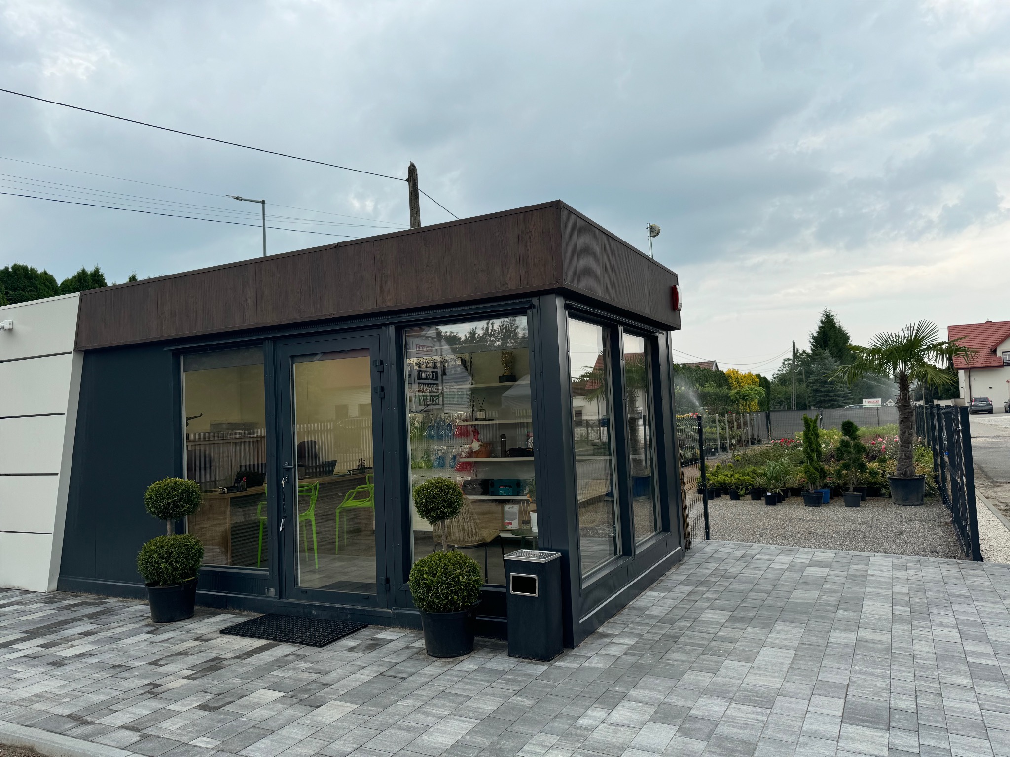 Nowoczesny pawilon sklepowy z dużymi oknami, prezentujący rośliny i akcesoria ogrodnicze, z kulistymi krzewami w donicach przed wejściem, wyłożony szarą kostką brukową.
