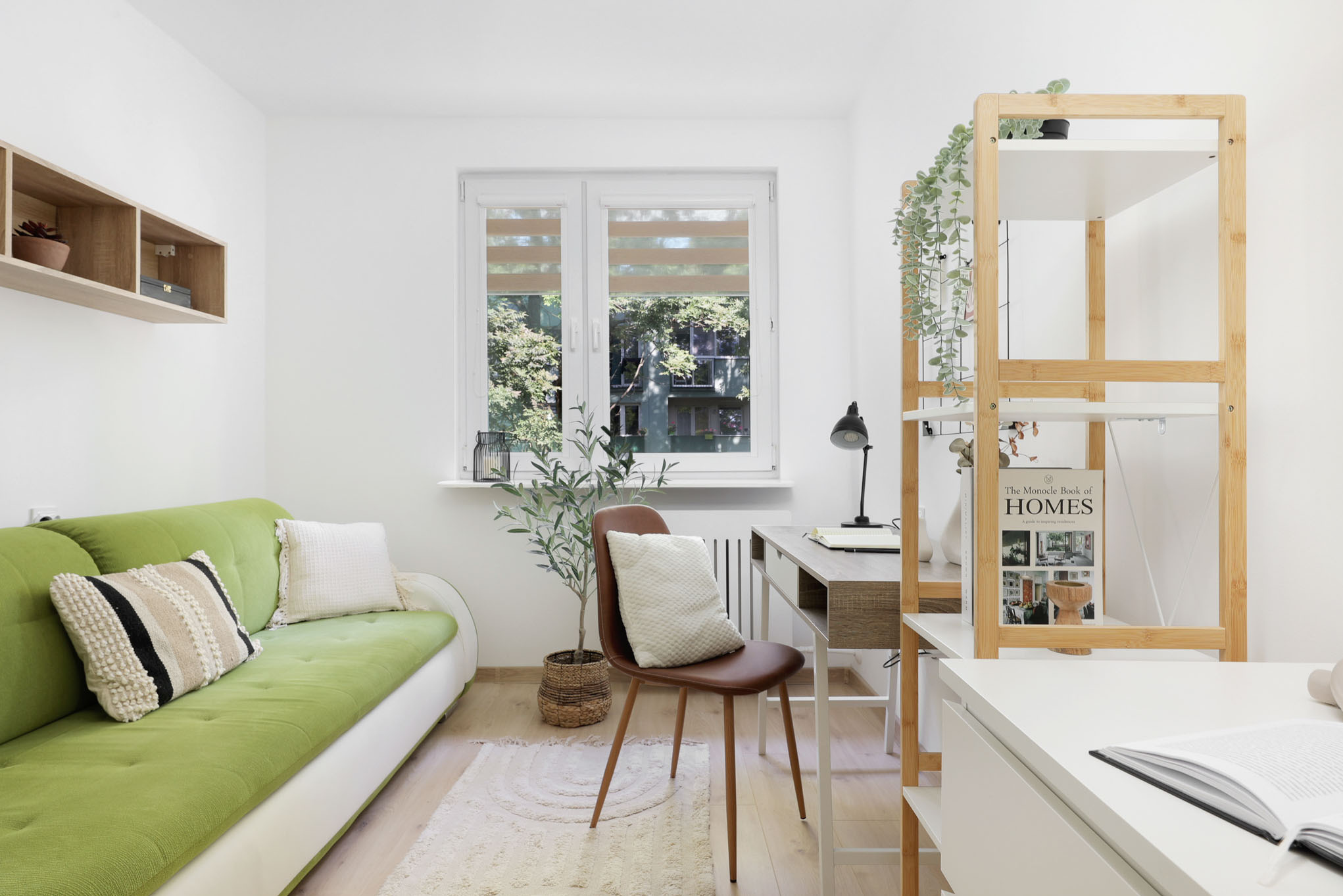 Jasne wnętrze pokoju z zieloną sofą, biurkiem, krzesłem i regałem, oświetlone naturalnym światłem z okna z roletami dzień-noc.