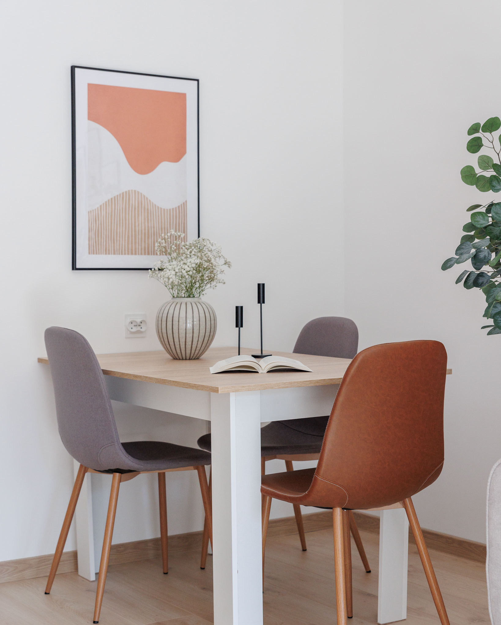 Jasny pokój z drewnianym stołem otoczonym szarymi i brązowym krzesłami, na blacie wazon z gipsówką, czarne świeczniki i otwarta książka, na ścianie abstrakcyjny obraz w czarnej ramie.