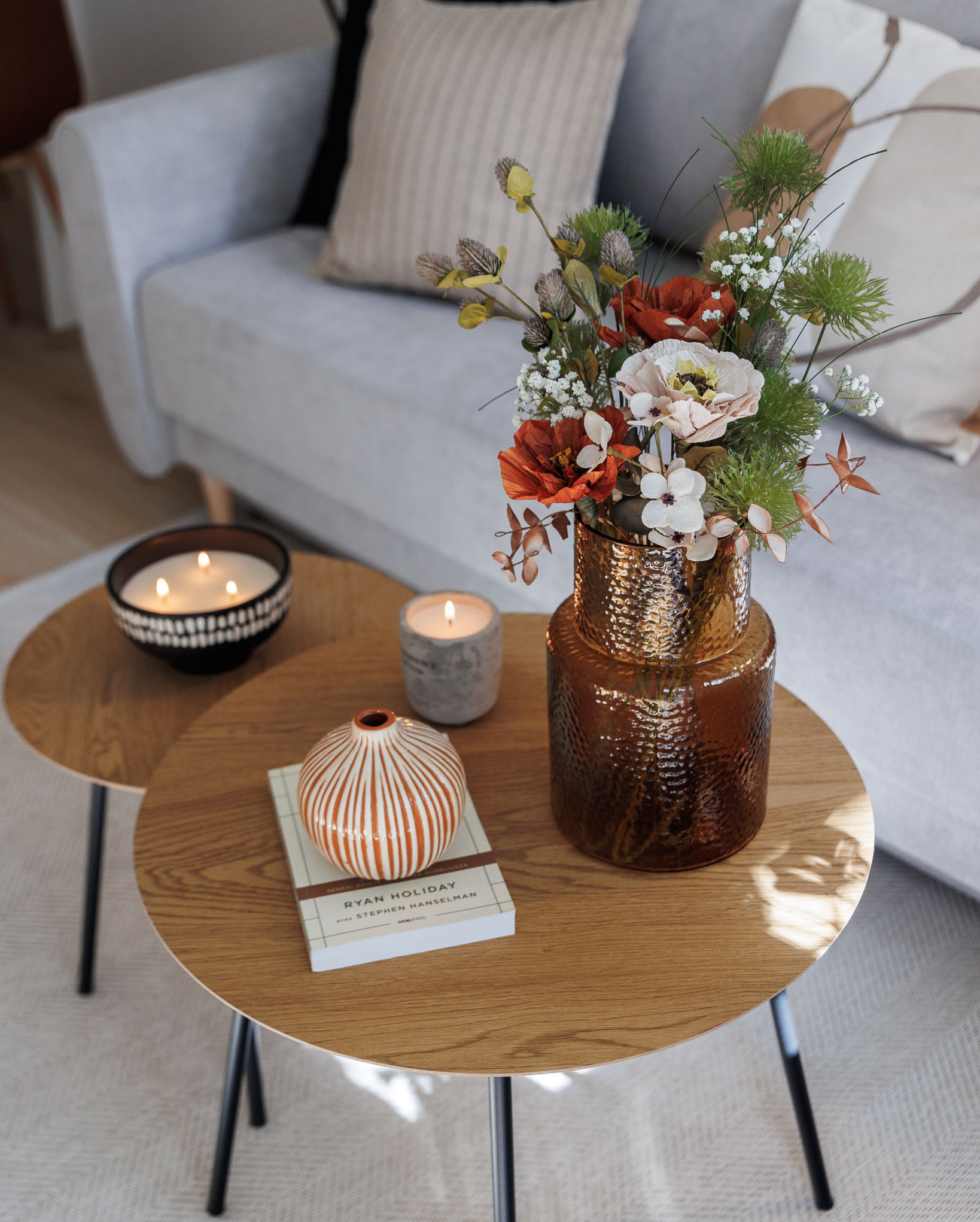 Aranżacja wnętrza: bukiet w brązowym, tłoczonym wazonie, mała prążkowana waza na książce Ryana Holiday'a, zapalone świece na okrągłych stolikach kawowych, szara sofa w tle.