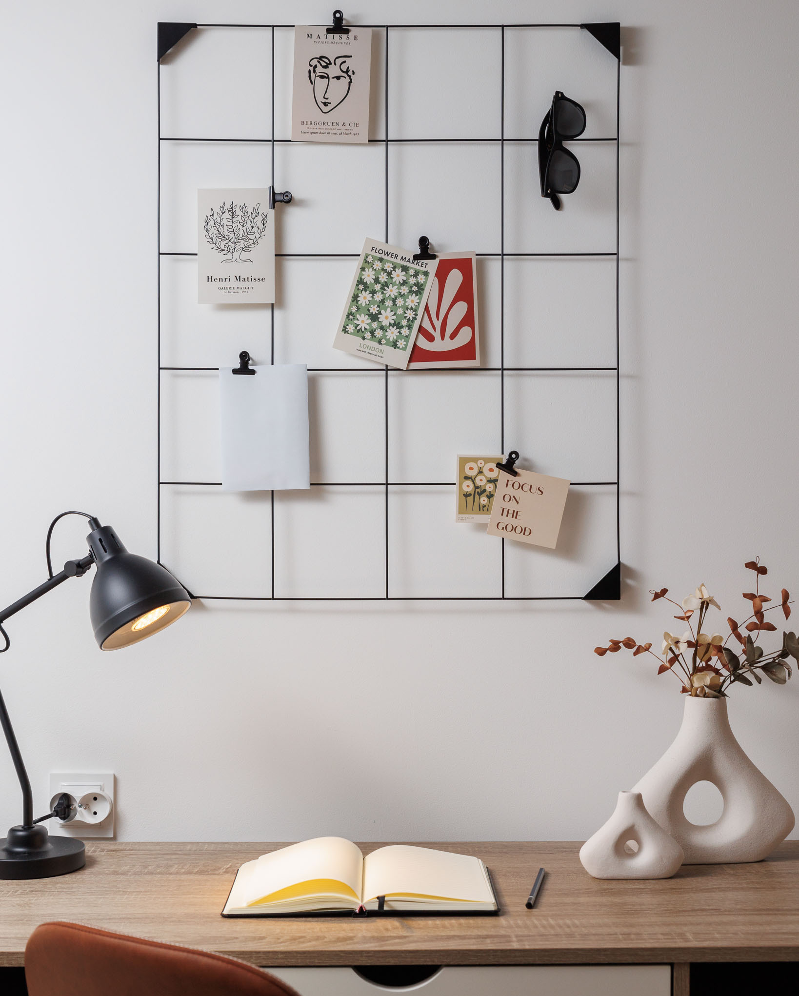 Biurko z otwartym notatnikiem, lampą i dekoracyjną ścianką z grafikami Matisse'a, wazonami ceramicznymi i okularami przeciwsłonecznymi.