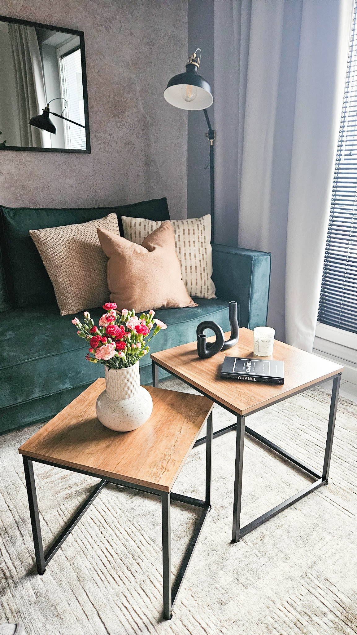 Stylowy salon z zieloną sofą, drewnianymi stolikami kawowymi z czarnymi nogami, wazonem z różowymi i czerwonymi goździkami, dekoracyjną rzeźbą, świecą i książką 'Chanel' na blacie. W tle zasłony...
