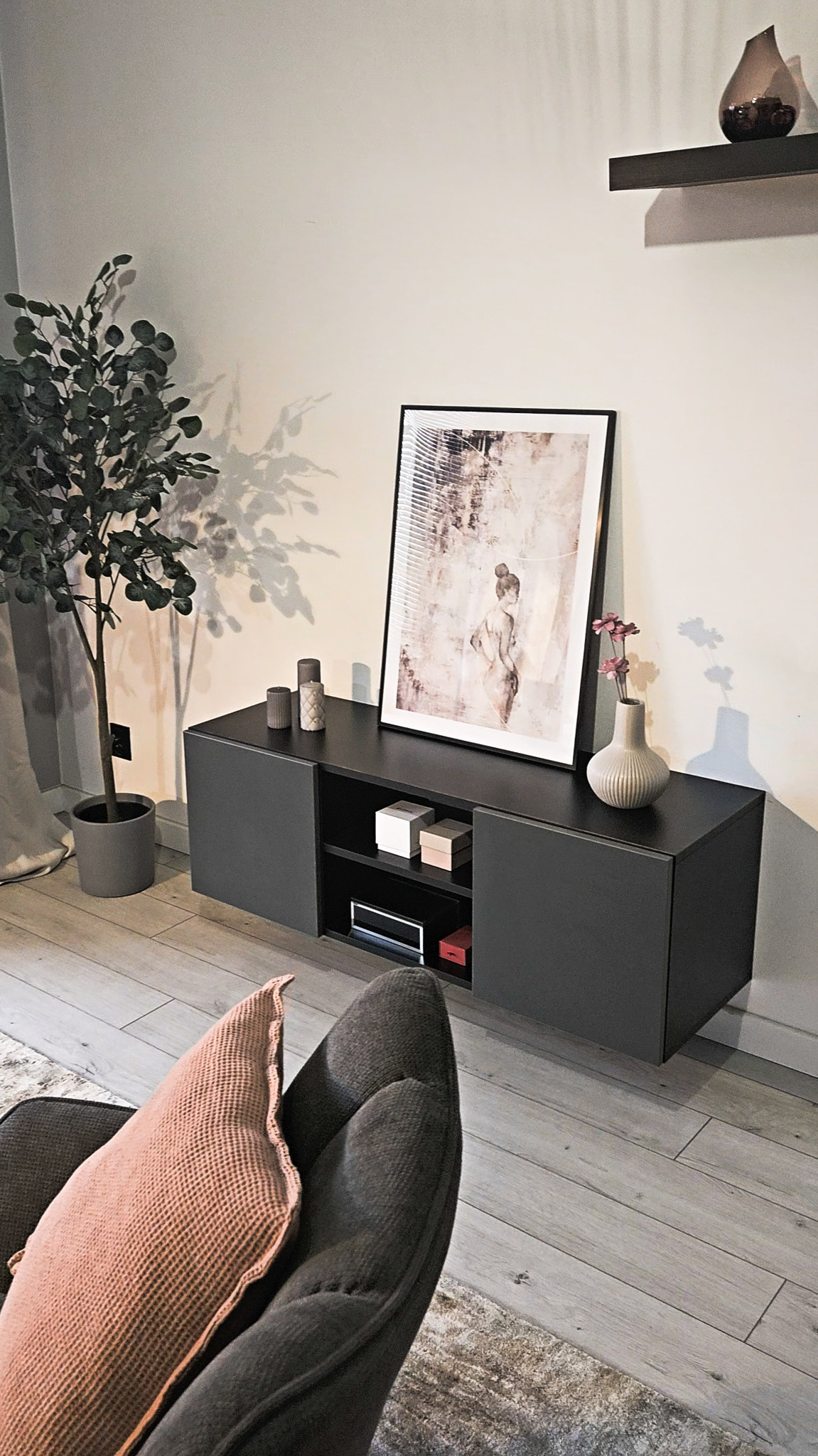 Nowoczesny salon z szarą szafką RTV, dekoracjami, obrazem w czarnej ramie, rośliną w doniczce i drewnianą podłogą w odcieniach szarości, widziany z perspektywy fotela.