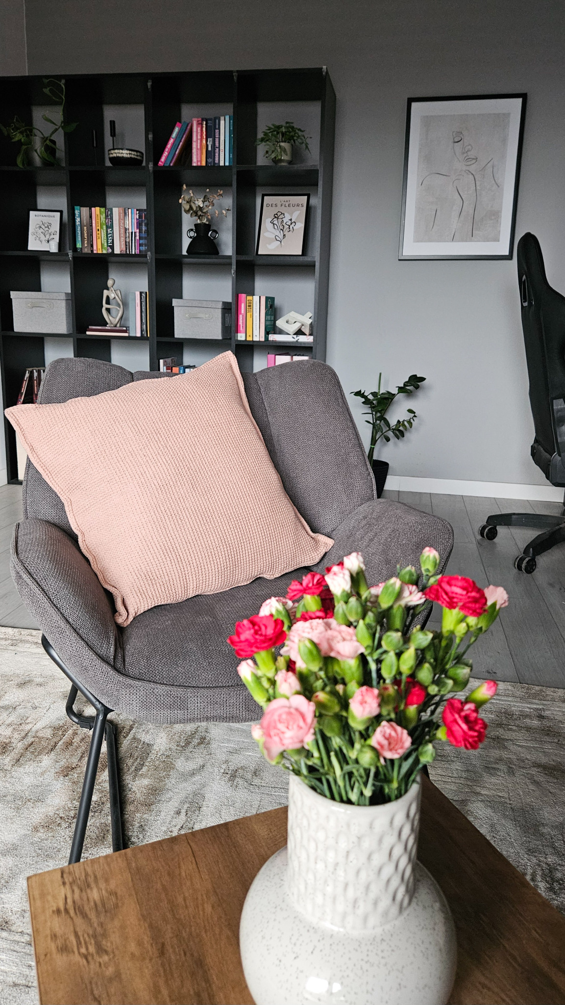 Szary fotel z różową poduszką i bukietem różowych i czerwonych goździków w białym wazonie na drewnianym stoliku, w tle regał z książkami i dekoracjami, minimalistyczny plakat na ścianie.