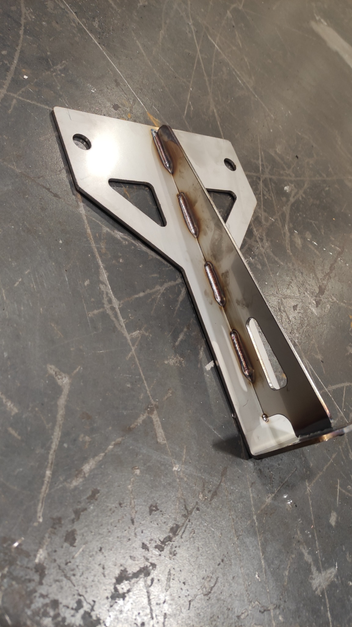 Spawany element konstrukcyjny z blachy stalowej na szorstkim tle warsztatowym, widoczne spoiny i otwory montażowe.