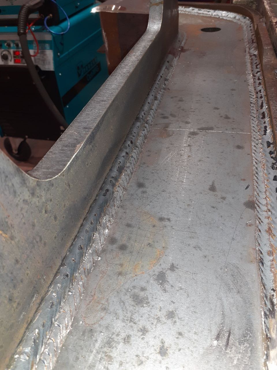 Spoina łącząca elementy metalowe, widoczna maszyna spawalnicza w tle.