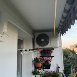 Klimatyzator na balkonie z kwiatami w doniczkach, widok na zieleń i budynki w oddali, oświetlenie balkonowe w postaci girlandy żarówkowej.