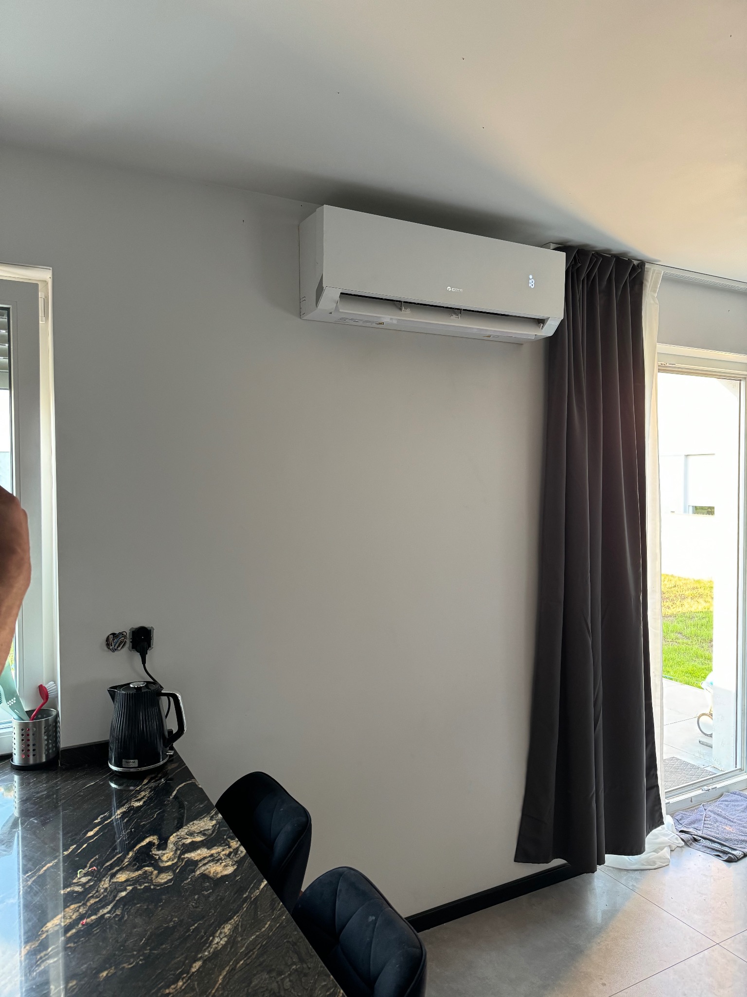 Biała jednostka klimatyzacji marki Gree zamontowana na ścianie w nowoczesnym wnętrzu, widoczny czarny czajnik elektryczny na blacie z marmuru i zasłony w oknie.
