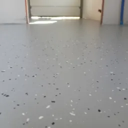 Jasnoszara posadzka z żywicy epoksydowej w garażu, z widocznymi drobnymi, czarnymi i białymi elementami dekoracyjnymi. Widok na otwartą bramę garażową.
