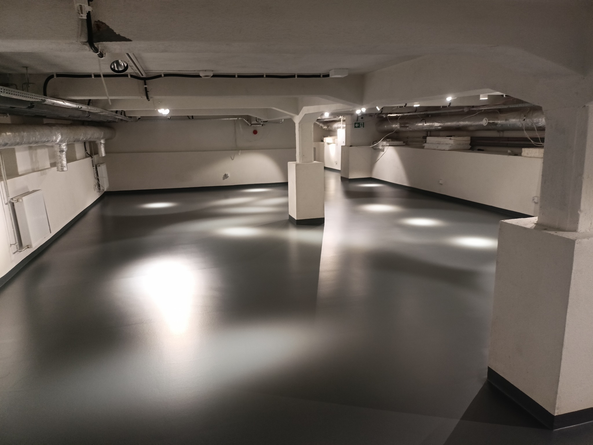 Błyszcząca, szara posadzka w przestronnym, industrialnym pomieszczeniu z betonowymi kolumnami i widocznymi instalacjami pod sufitem. Światło odbija się od gładkiej powierzchni podłogi.