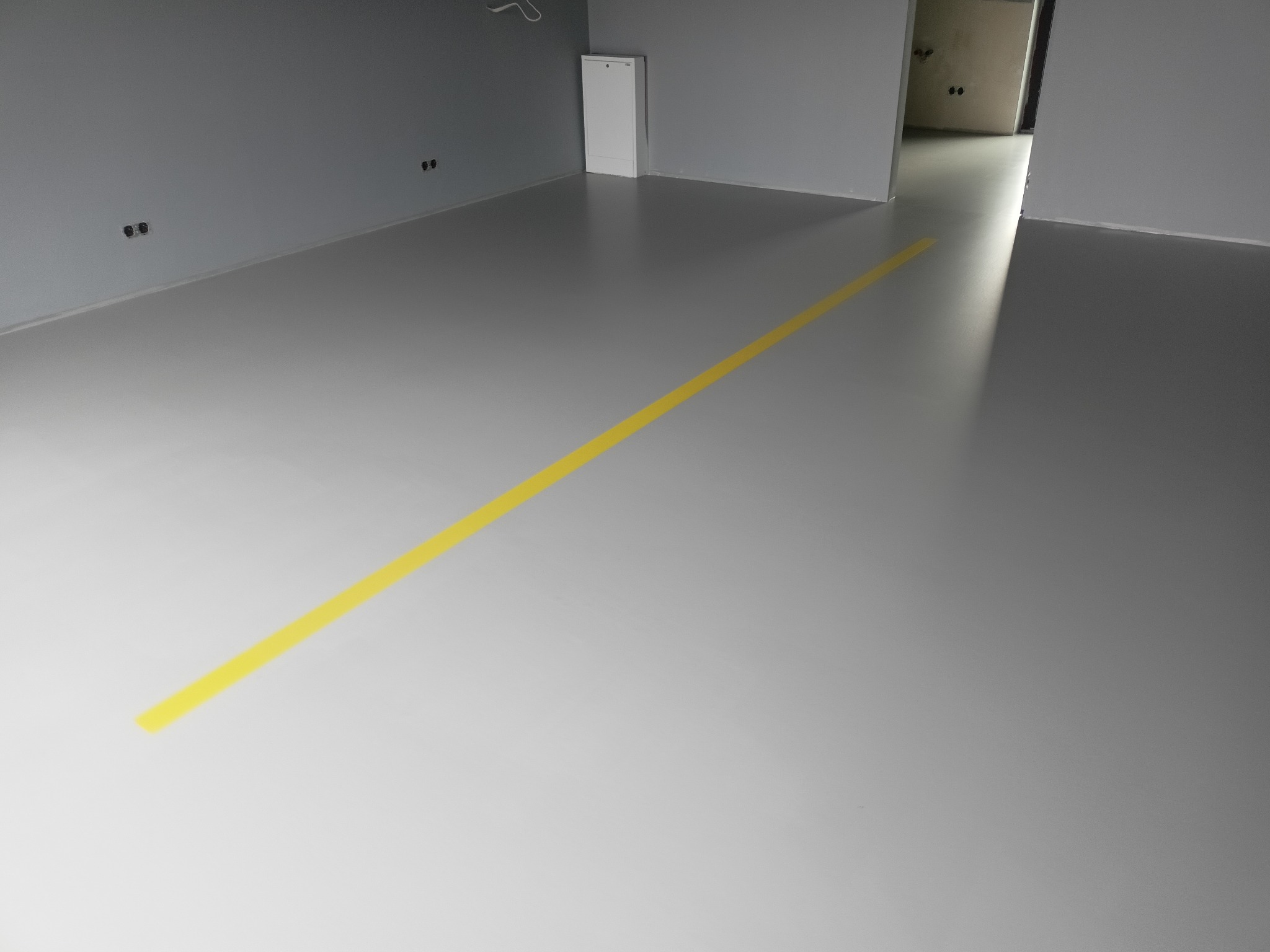 Szara posadzka w surowym wnętrzu, oznaczona żółtą linią, widoczne puszki elektryczne na ścianie i drzwi do kolejnego pomieszczenia.