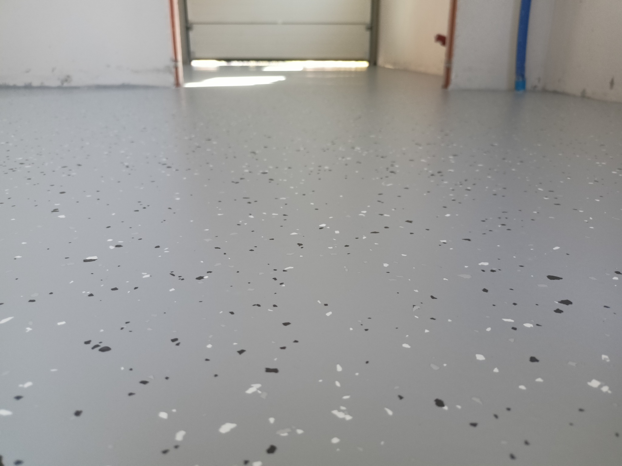 Jasnoszara posadzka z żywicy epoksydowej w garażu, z widocznymi drobnymi, czarnymi i białymi elementami dekoracyjnymi. Widok na otwartą bramę garażową.