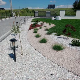 Kaskadowy ogród z białego żwiru, ozdobnego krzewu i lamp ogrodowych, widok na bramę i dom w tle.